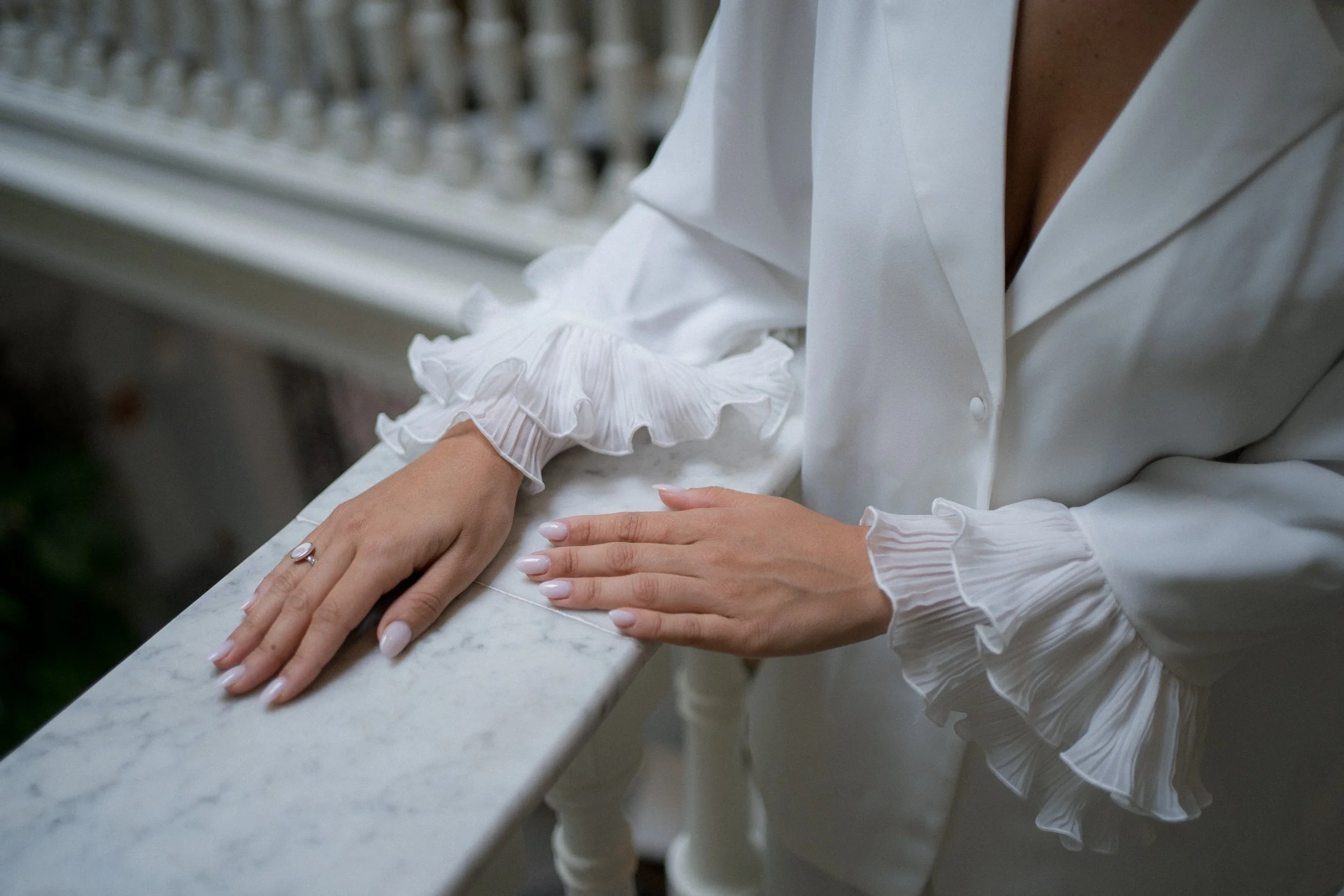 Manos con uñas arregladas y anillo, descansando sobre una mesa de mármol, con blusa blanca con mangas con volantes rizados.