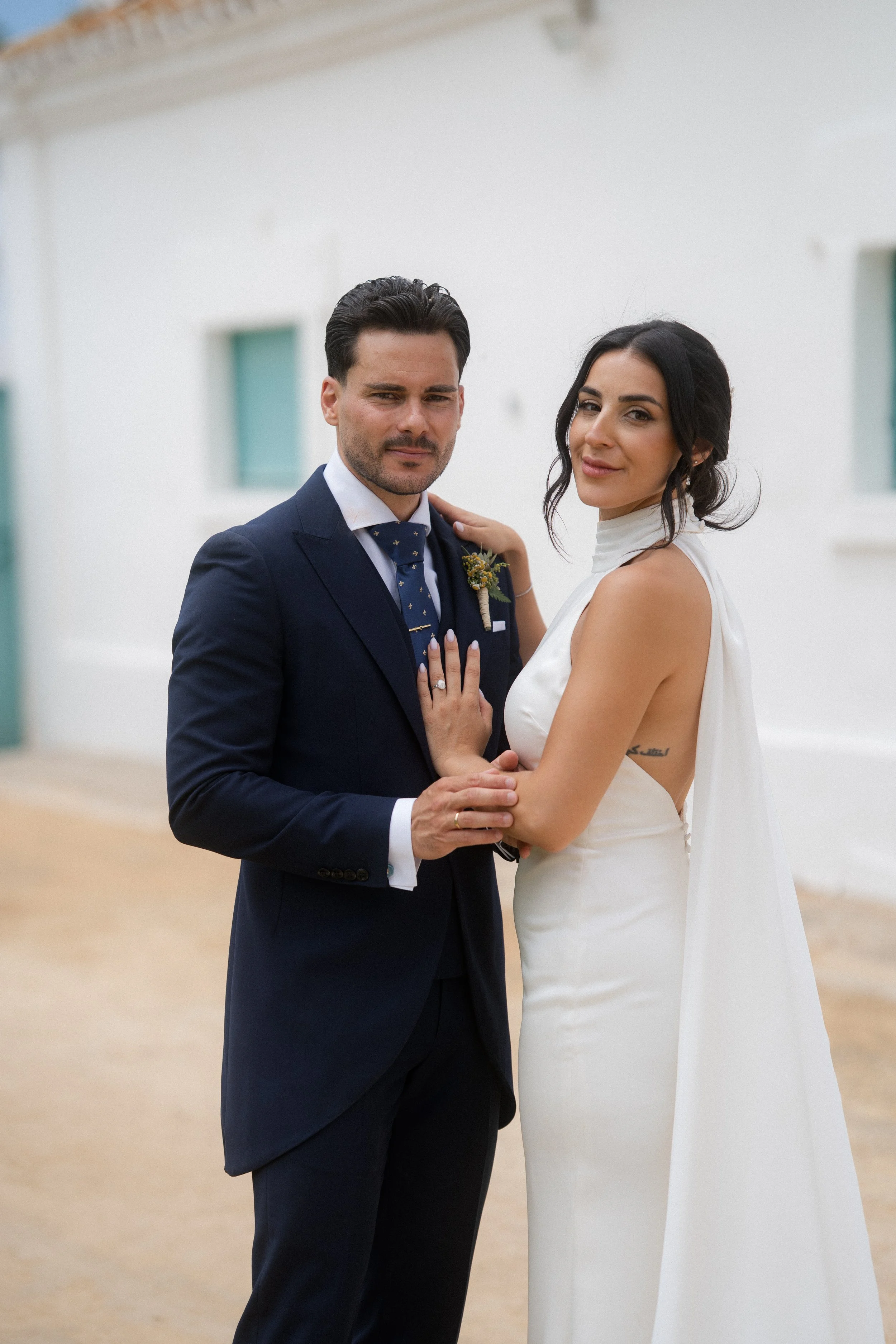 Pareja en traje de novia y novio en un entorno interior con paredes blancas y ventanas azules.