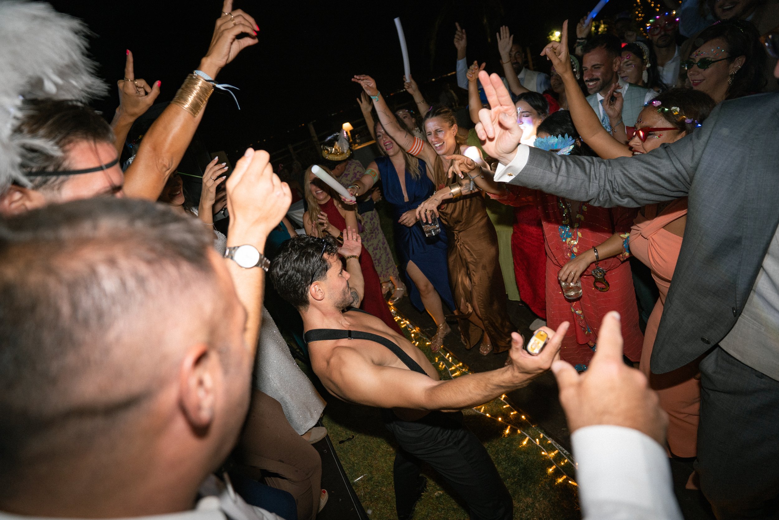 Personas bailando y disfrutando en una fiesta elegante al aire libre por la noche, con luces decorativas y gente vestida con ropa formal y colorida.
