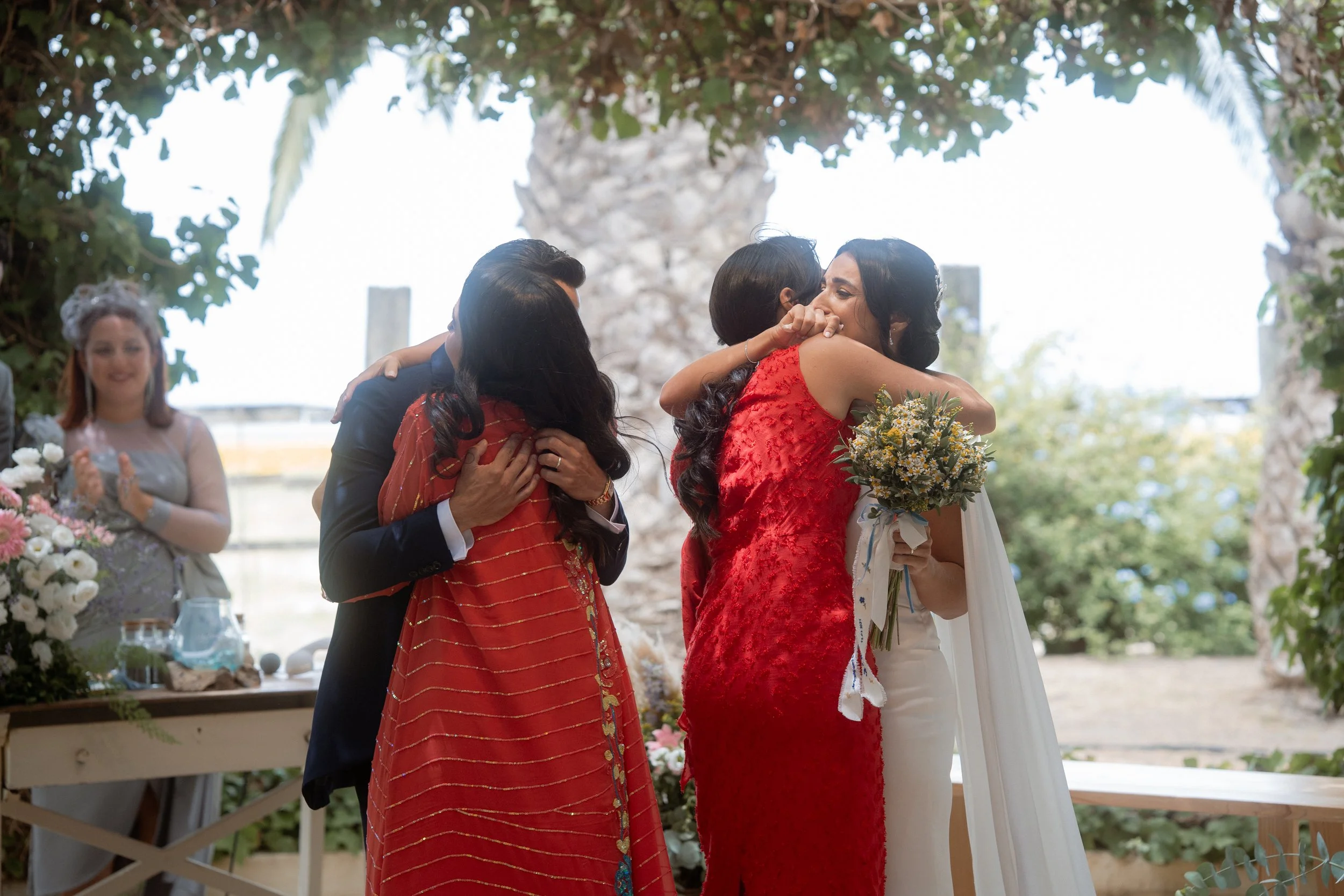 Una boda al aire libre donde dos mujeres en vestidos rojos se abrazan emocionadas y una de ellas sostiene un ramo de flores. En el fondo, una mujer observa y a la izquierda hay mesas con decoraciones florales.