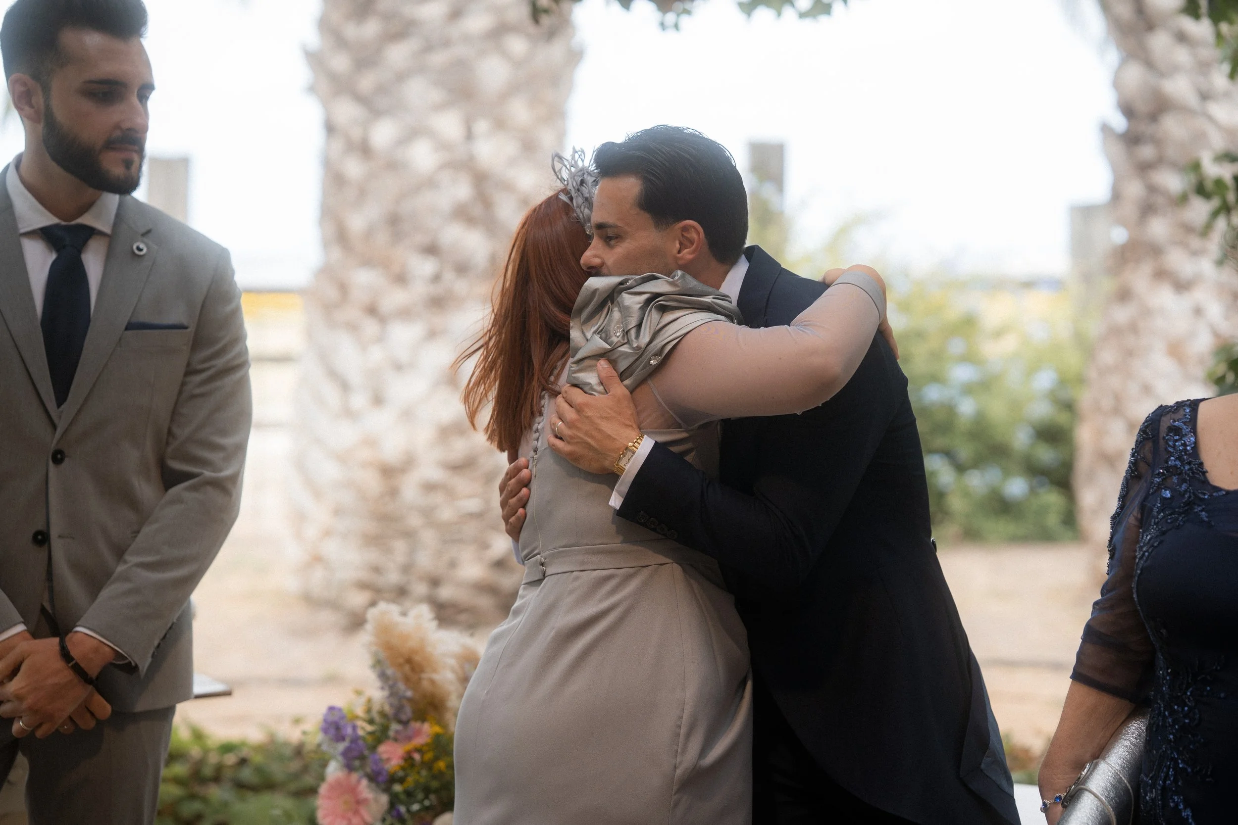 Una pareja abrazándose con cariño en una ceremonia o evento formal, con otros asistentes observando, en un entorno al aire libre con columnas de piedra y flores.