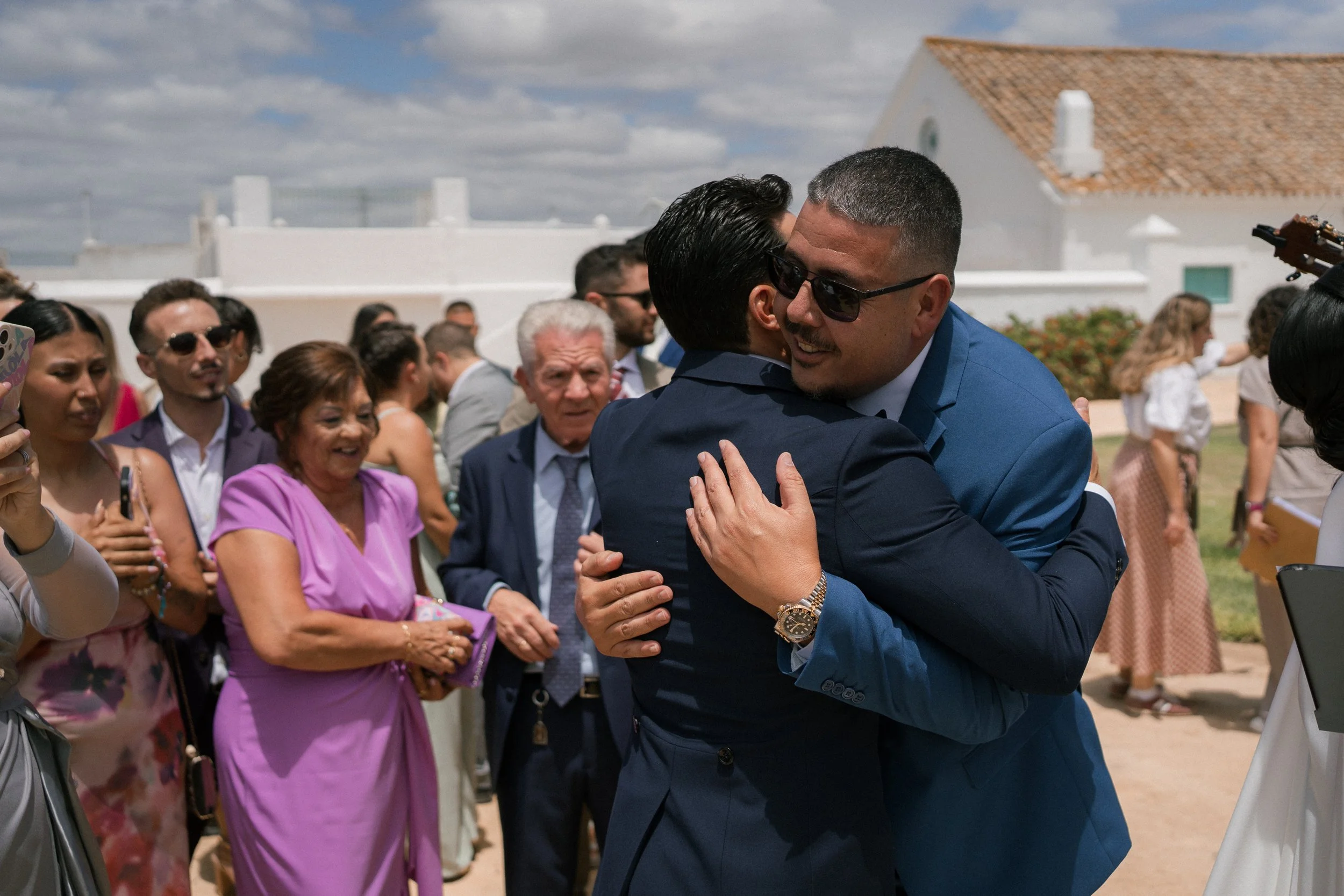 Dos hombres en trajes de negocios abrazándose en una celebración al aire libre, rodeados de varias personas que observan y disfrutan del momento.