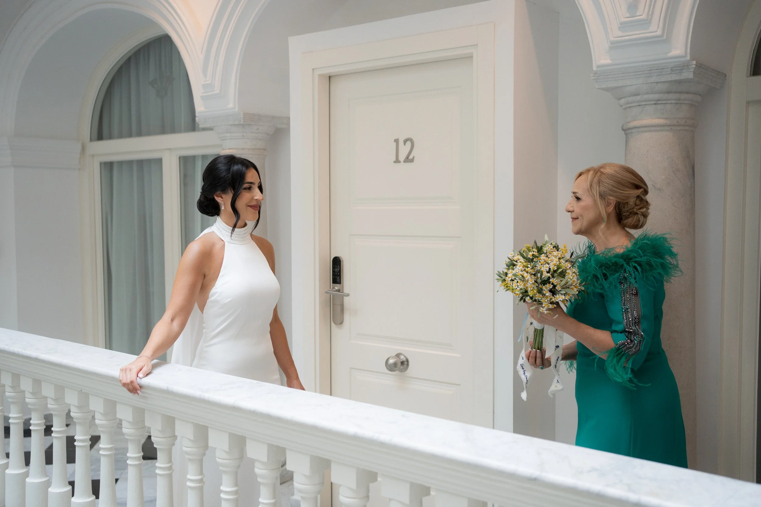 Mujer joven en vestido blanco mirando a mujer mayor en vestido verde que le ofrece un ramo de flores, en un pasillo elegante con columnas y puerta con número 12.