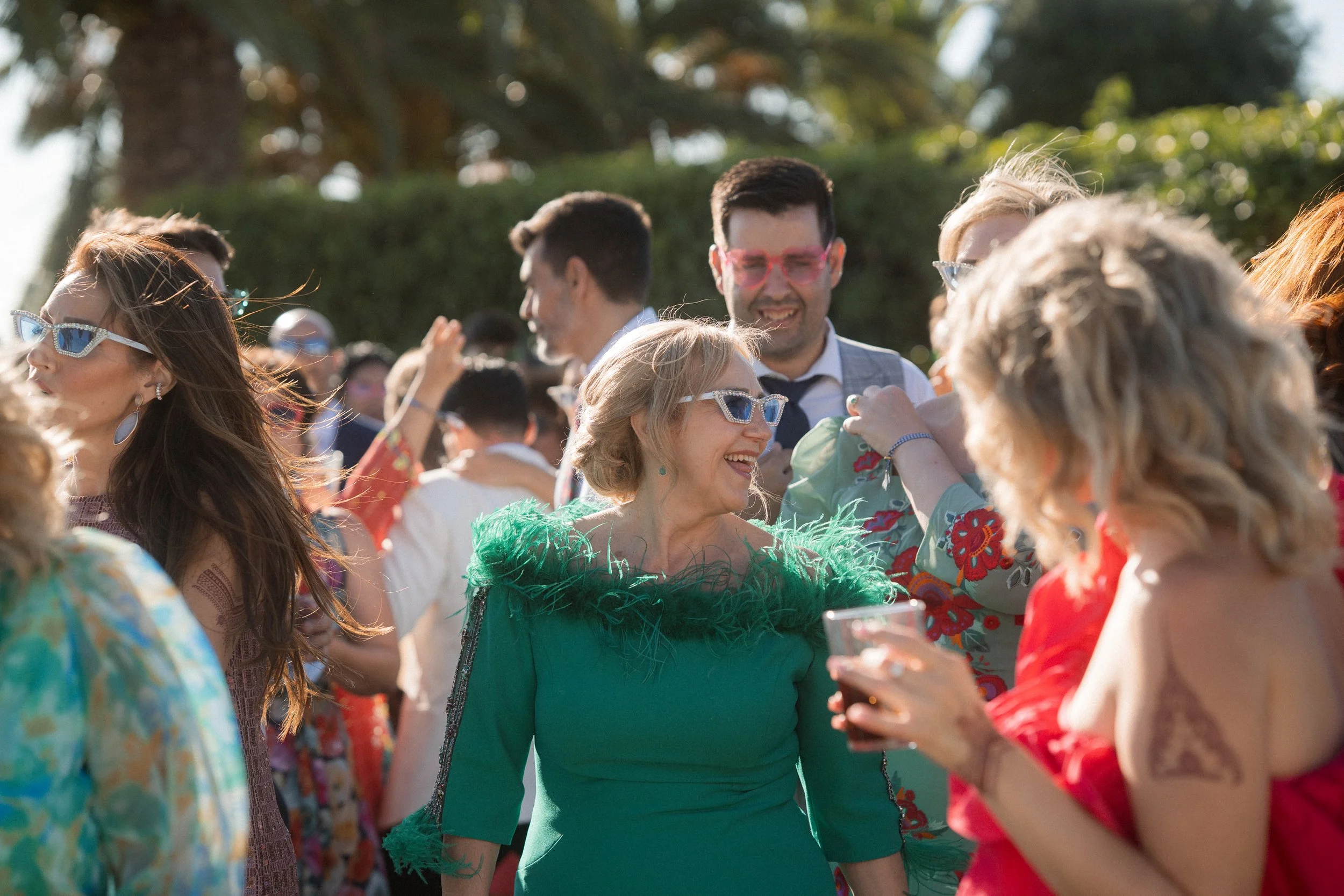 Grupo de personas en una boda al aire libre, disfrutando, algunas con gafas de sol y ropa colorida, en día soleado.