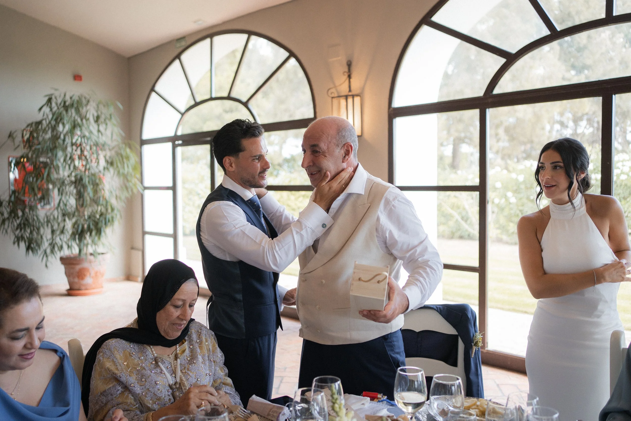 Hombre mayor recibe un regalo de un hombre más joven en una celebración en un comedor con grandes ventanas y otras personas observando, en un ambiente elegante y alegre.