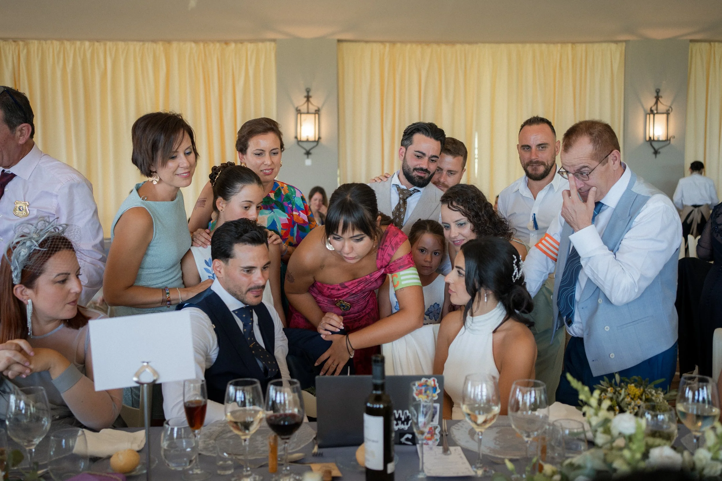Personas en una mesa observando una computadora portátil en una celebración, con varias copas de vino y decoración floral.