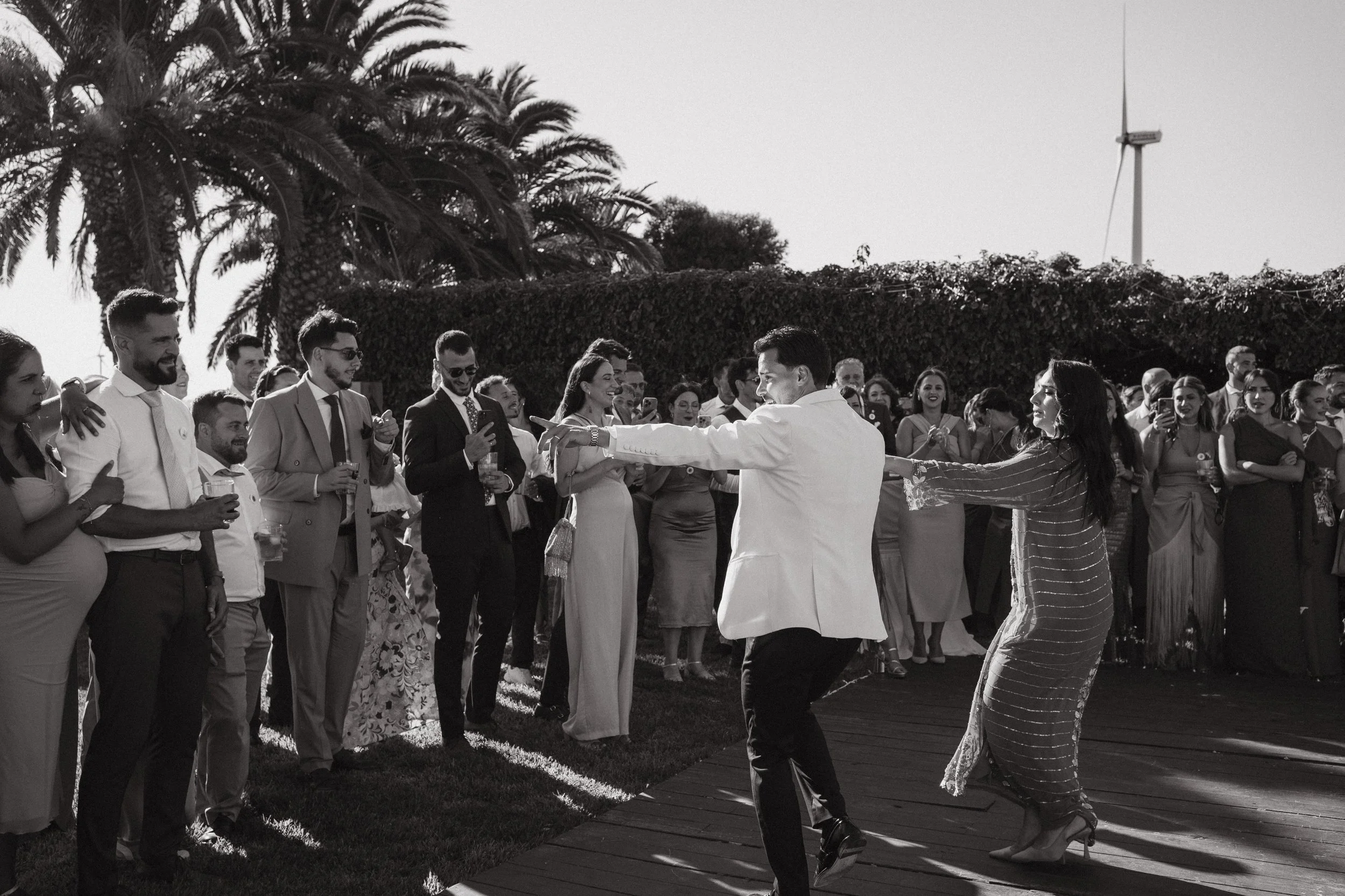 Pareja bailando en una boda, rodeados de invitados que disfrutan y toman fotos al aire libre.
