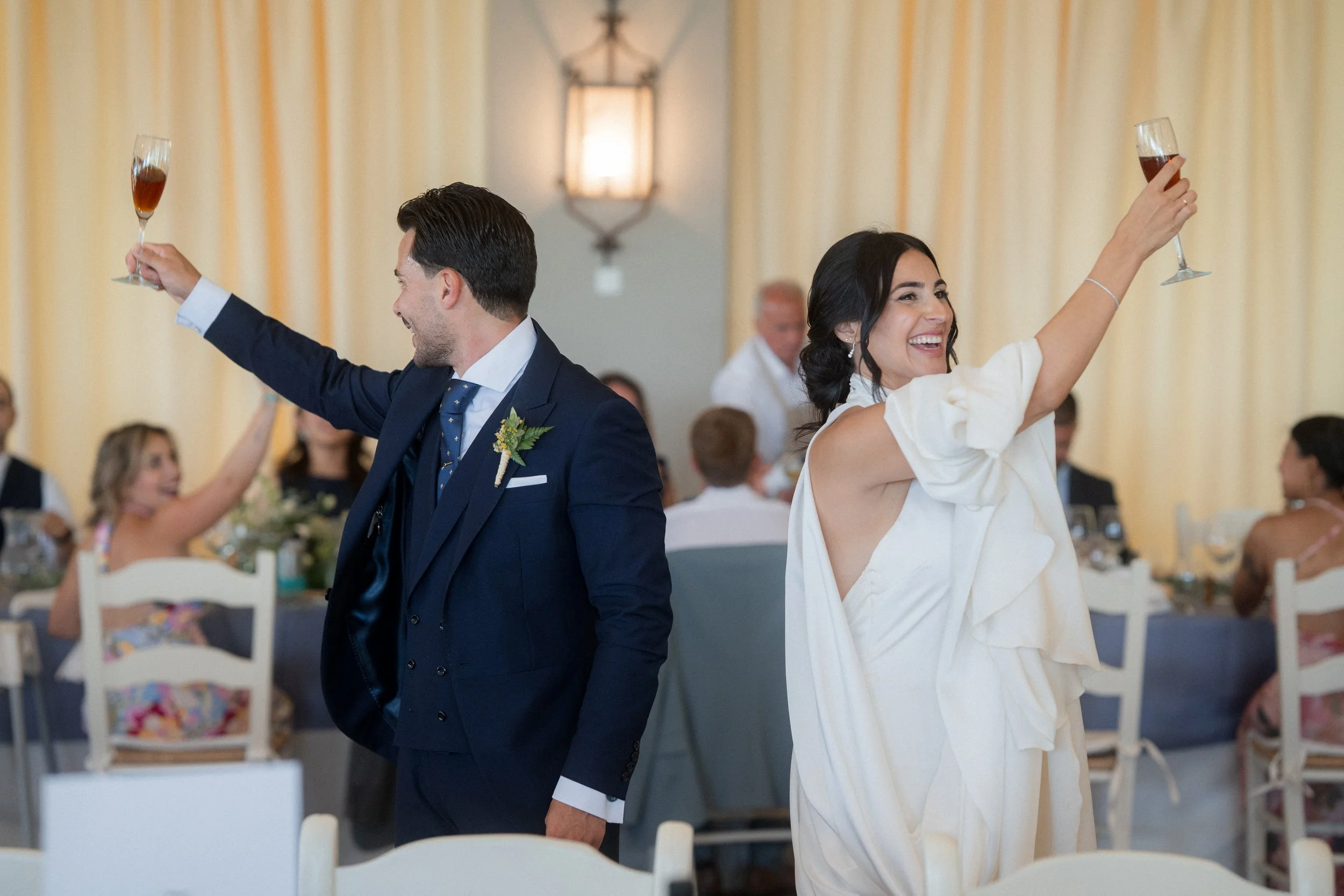 Una pareja feliz en una boda, sosteniendo copas de vino y sonriendo, en un salón decorado con cortinas y luces