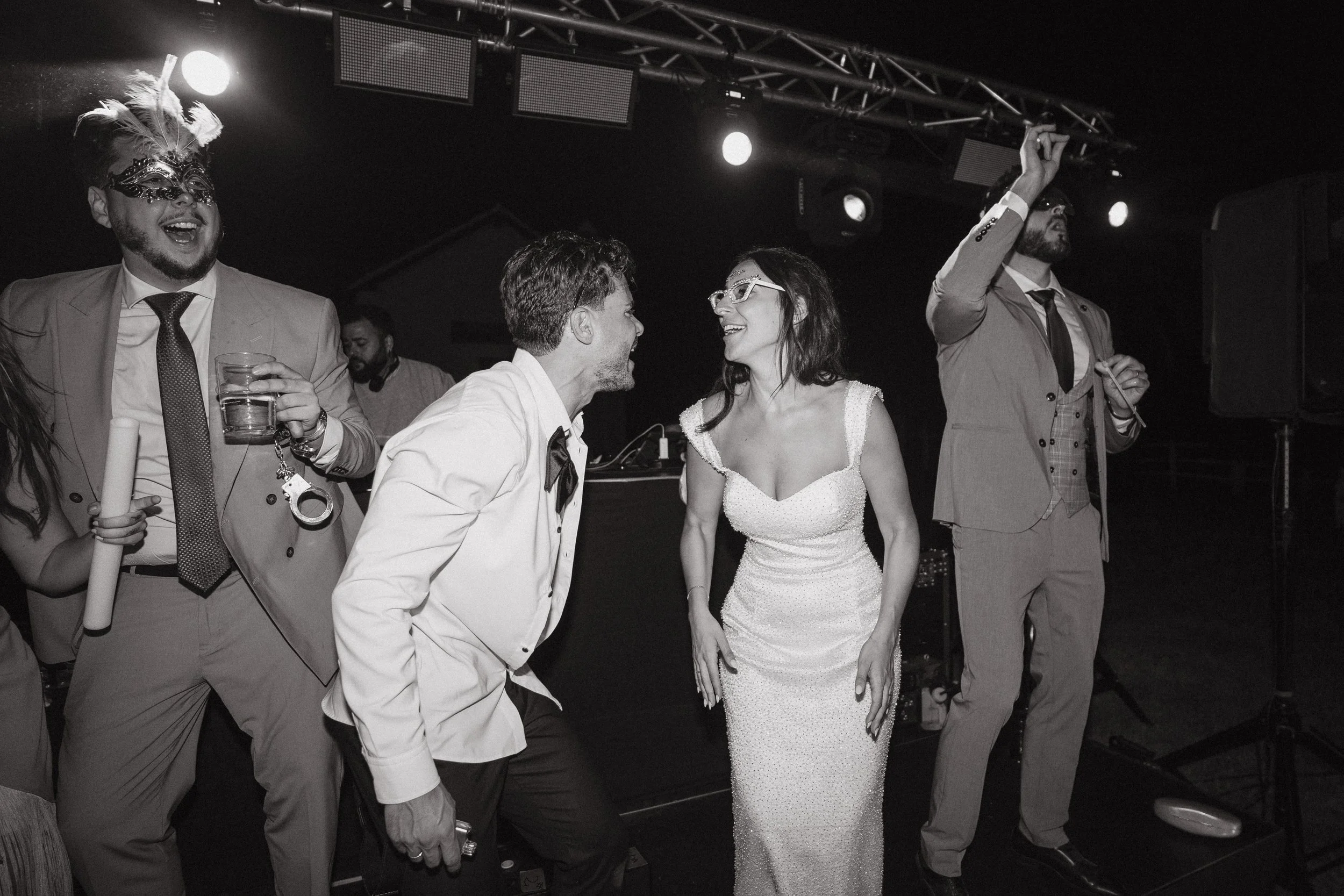 Grupo de personas en una fiesta, algunos vestidos formalmente, riendo y conversando, con luces de escenario y equipo de sonido al fondo.