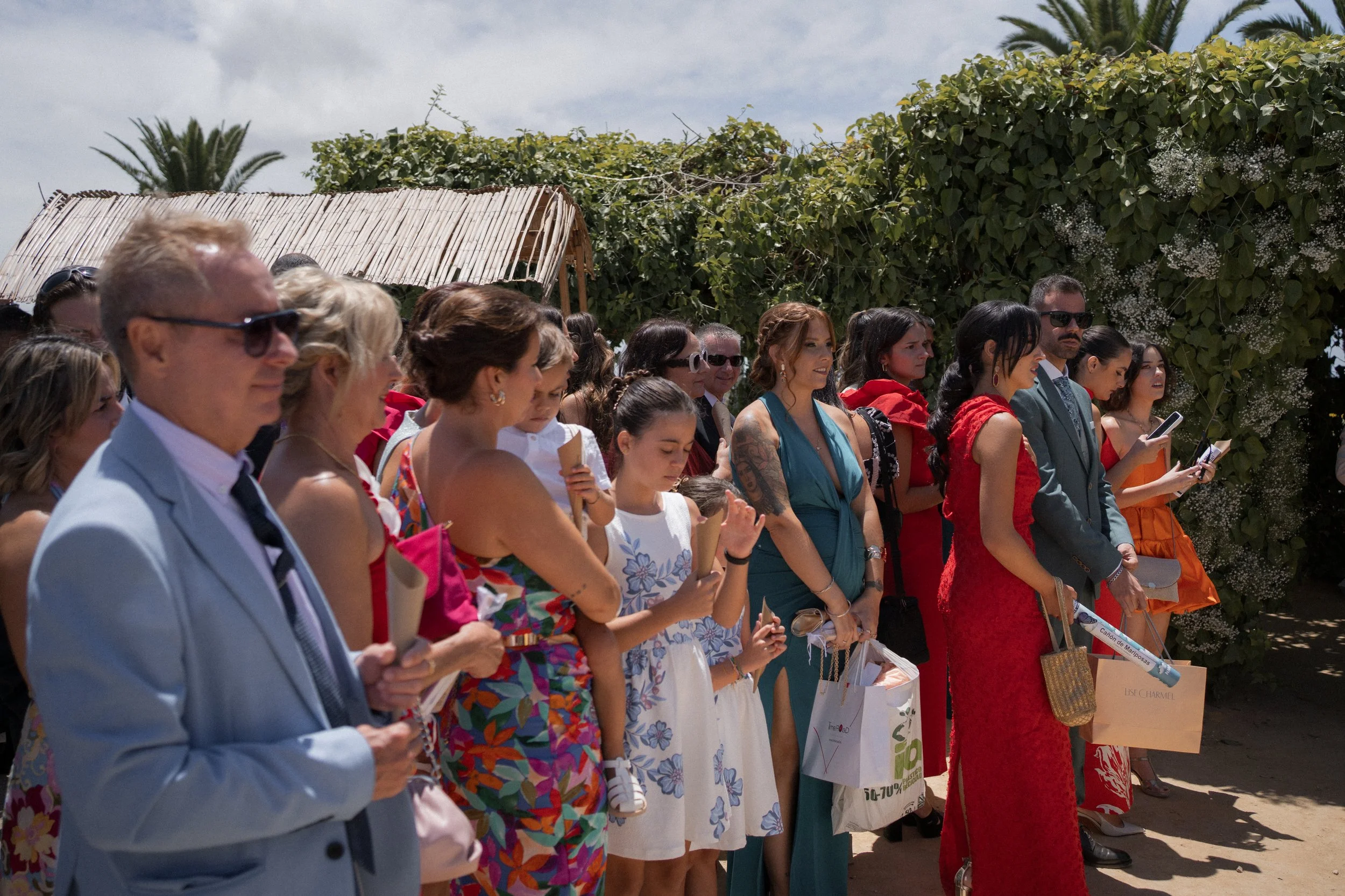 Grupo de personas en una ceremonia al aire libre, entre ellas mujeres con vestidos coloridos y hombres con trajes. La escena es bajo un cielo despejado con vegetación de fondo.