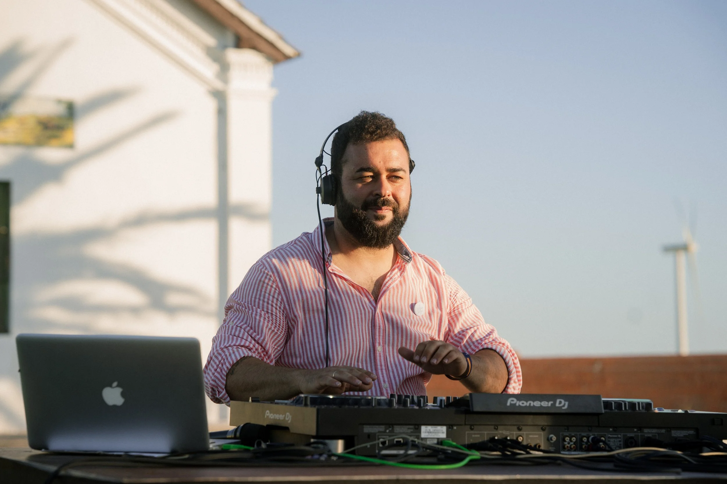 Un hombre con barba y camisa de rayas rojas y blancas, usando audífonos, mezclando música con equipo de DJ en una fiesta al aire libre durante el día.