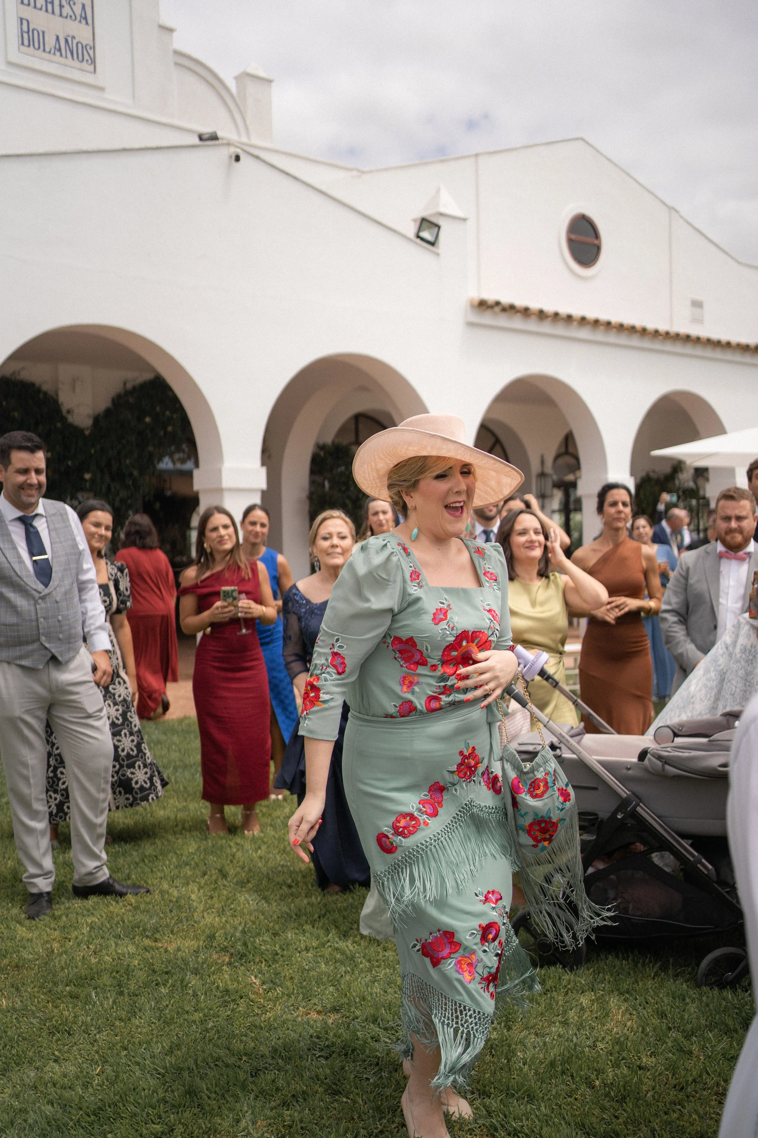 Mujer con sombrero y vestido bordado en un evento al aire libre rodeada de invitados.