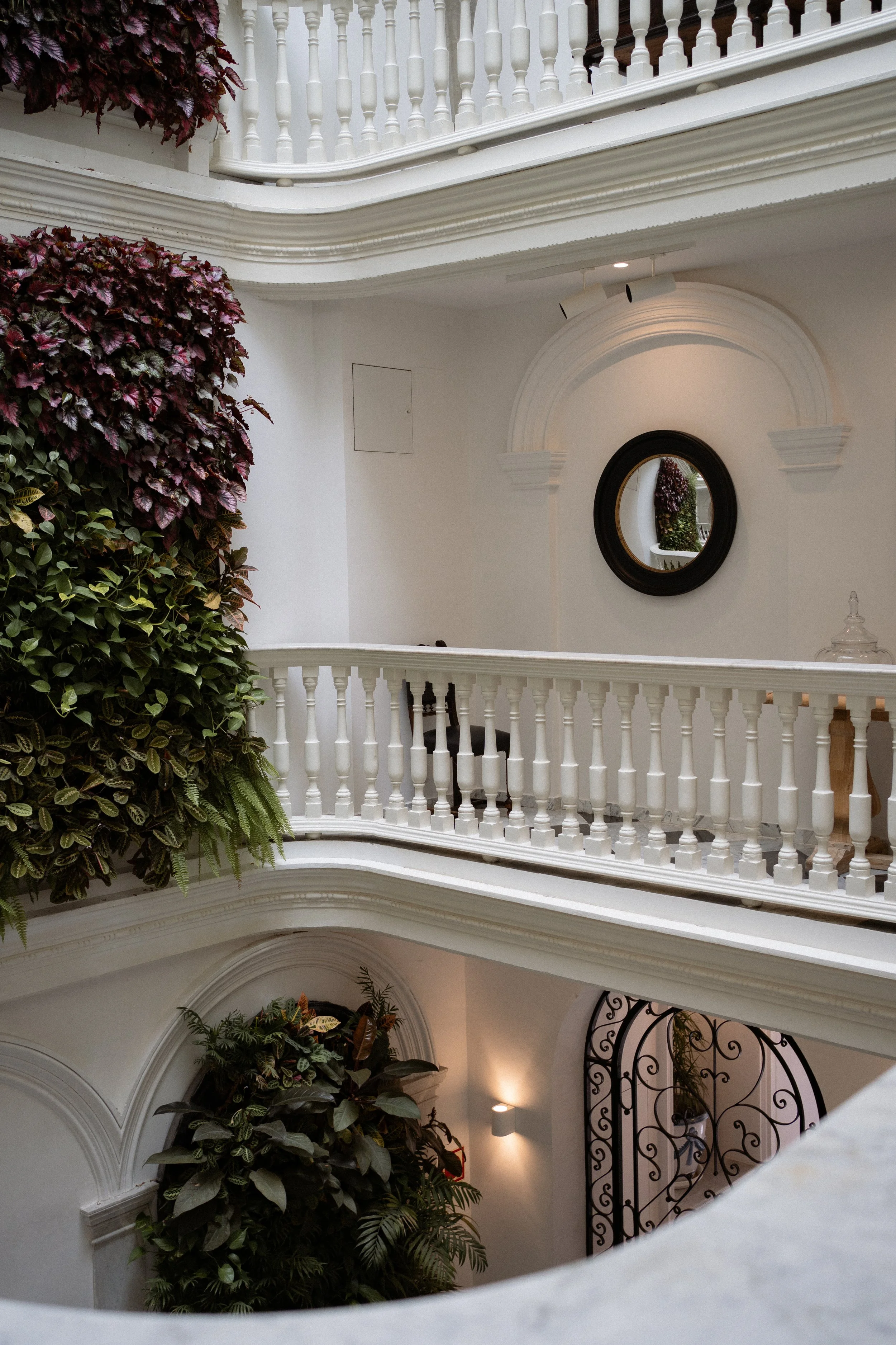 Interior de un edificio con pasarela blanca, plantas verdes y flores, espejo redondo en la pared y barandales blancos decorativos.
