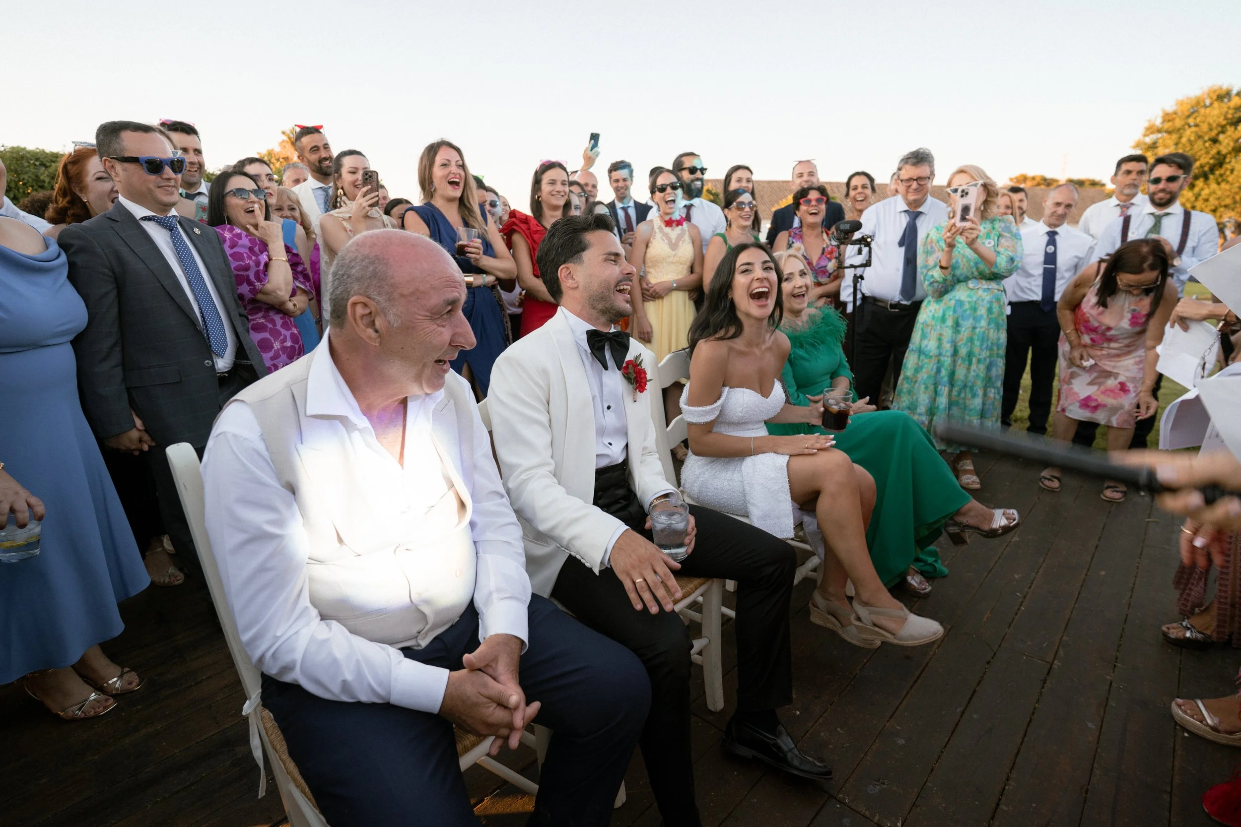 Grupo de personas riendo y celebrando en una boda al aire libre, algunos sentados y otros de pie, en un día soleado.