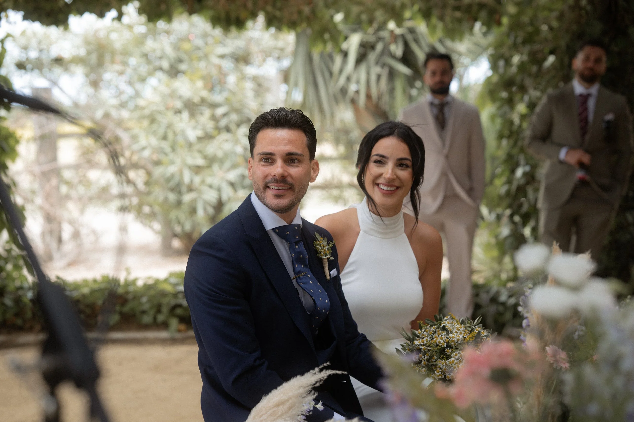 Pareja de novios en una boda en un entorno natural, la mujer con vestido blanco y el hombre con traje azul oscuro, rodeados de amigos y familiares.