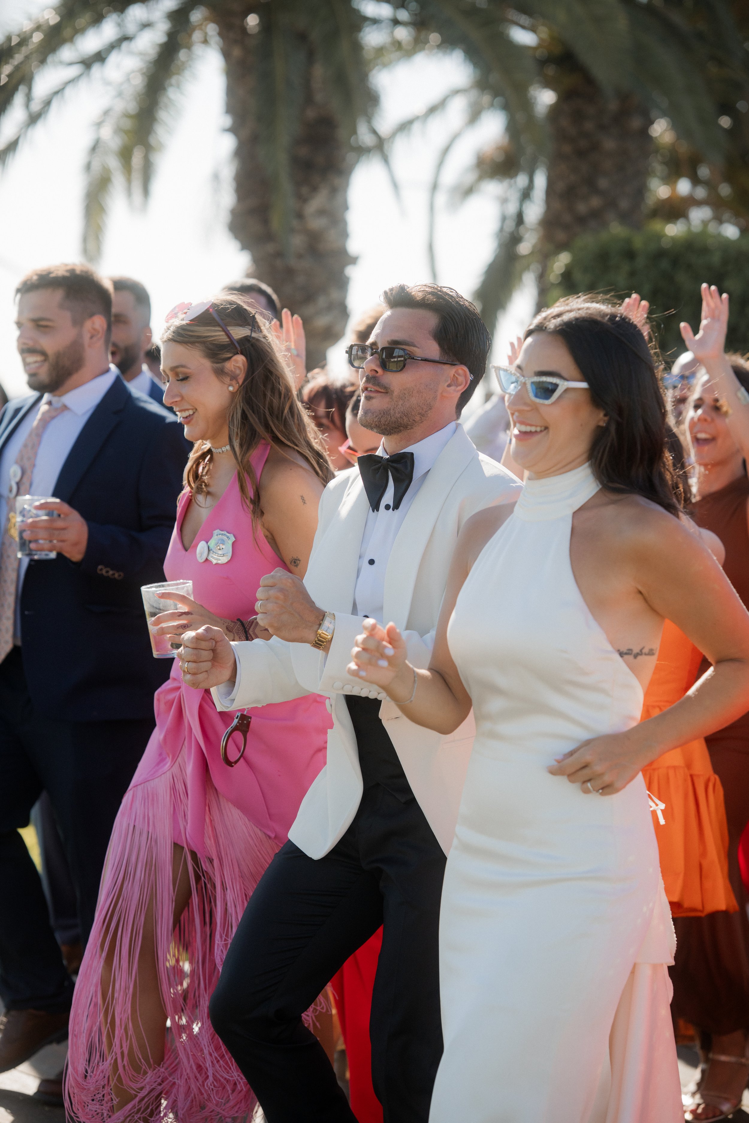 Grupo de personas bailando en una fiesta al aire libre, con árboles y palmas en el fondo, algunos llevan gafas y están sonriendo.