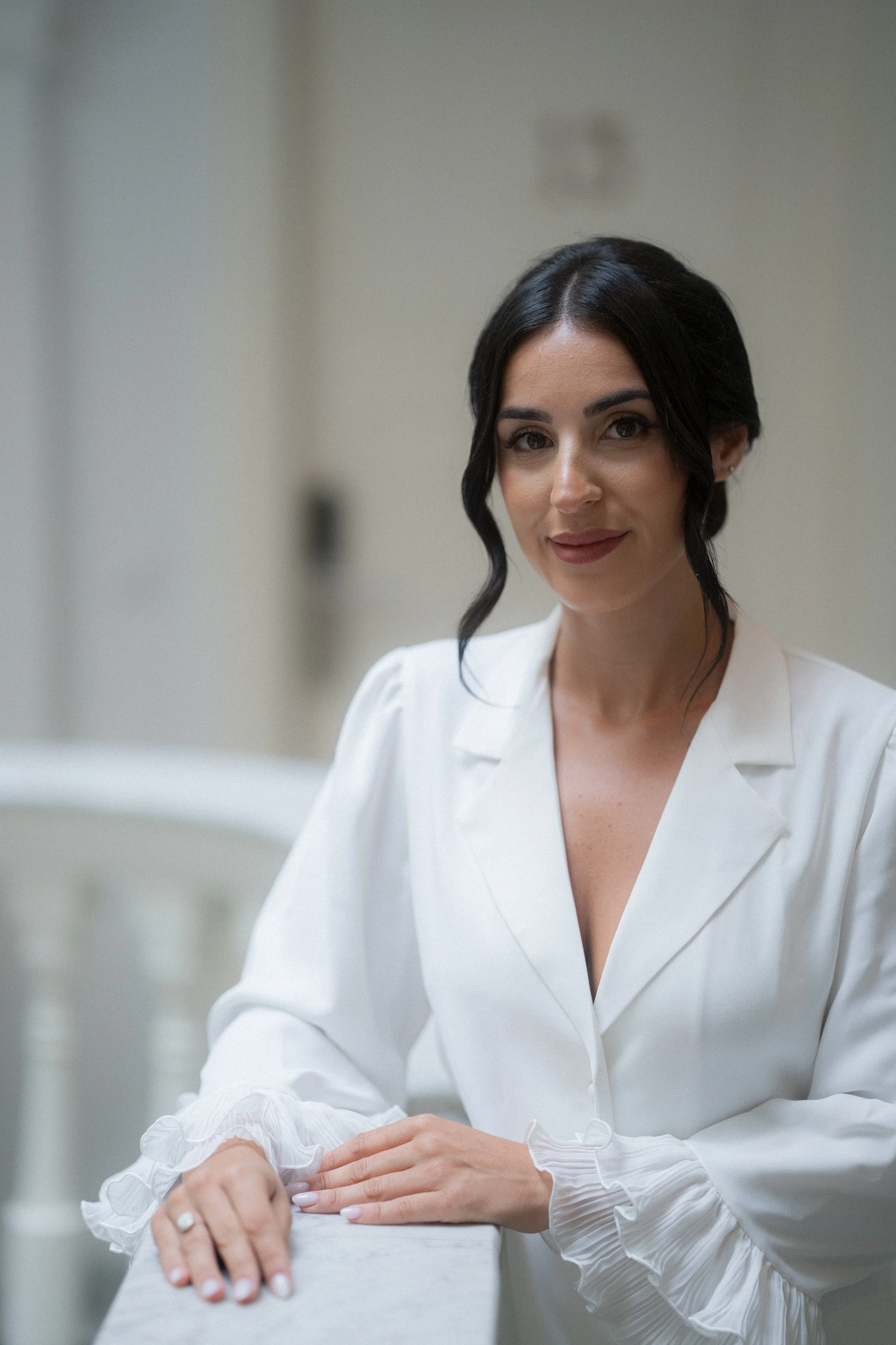 Mujer con cabello oscuro y rizado, vestida con blusa blanca, sonriendo y posando en un entorno interior.
