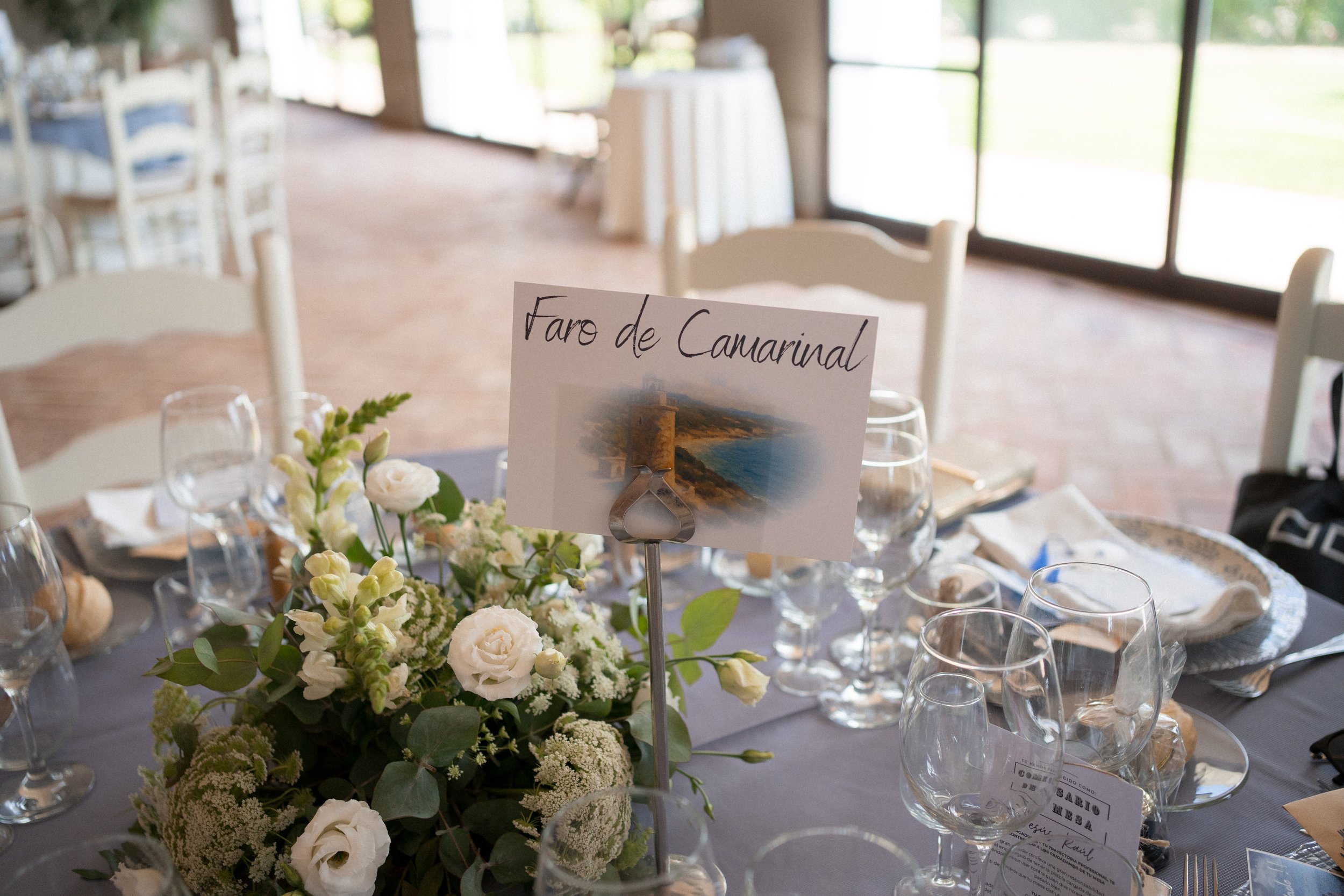Mesa decorada con flores blancas y cristalería, con señal que dice "Faro de Camarinal" y una foto del faro, en un salón con ventanas grandes y vista al exterior.