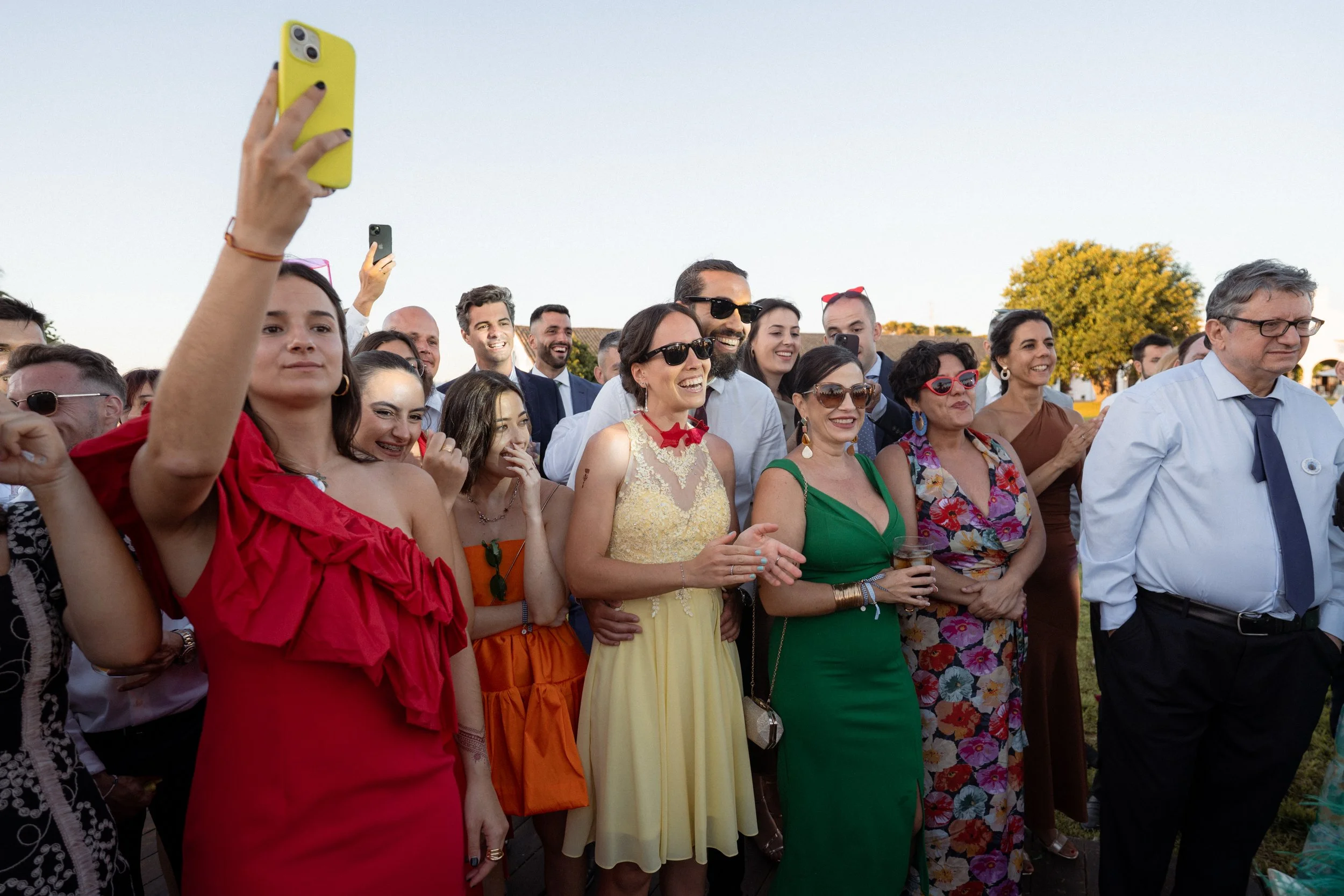 Grupo de personas en una reunión al aire libre, sonriendo y celebrando, con varias personas tomando fotos con sus teléfonos.