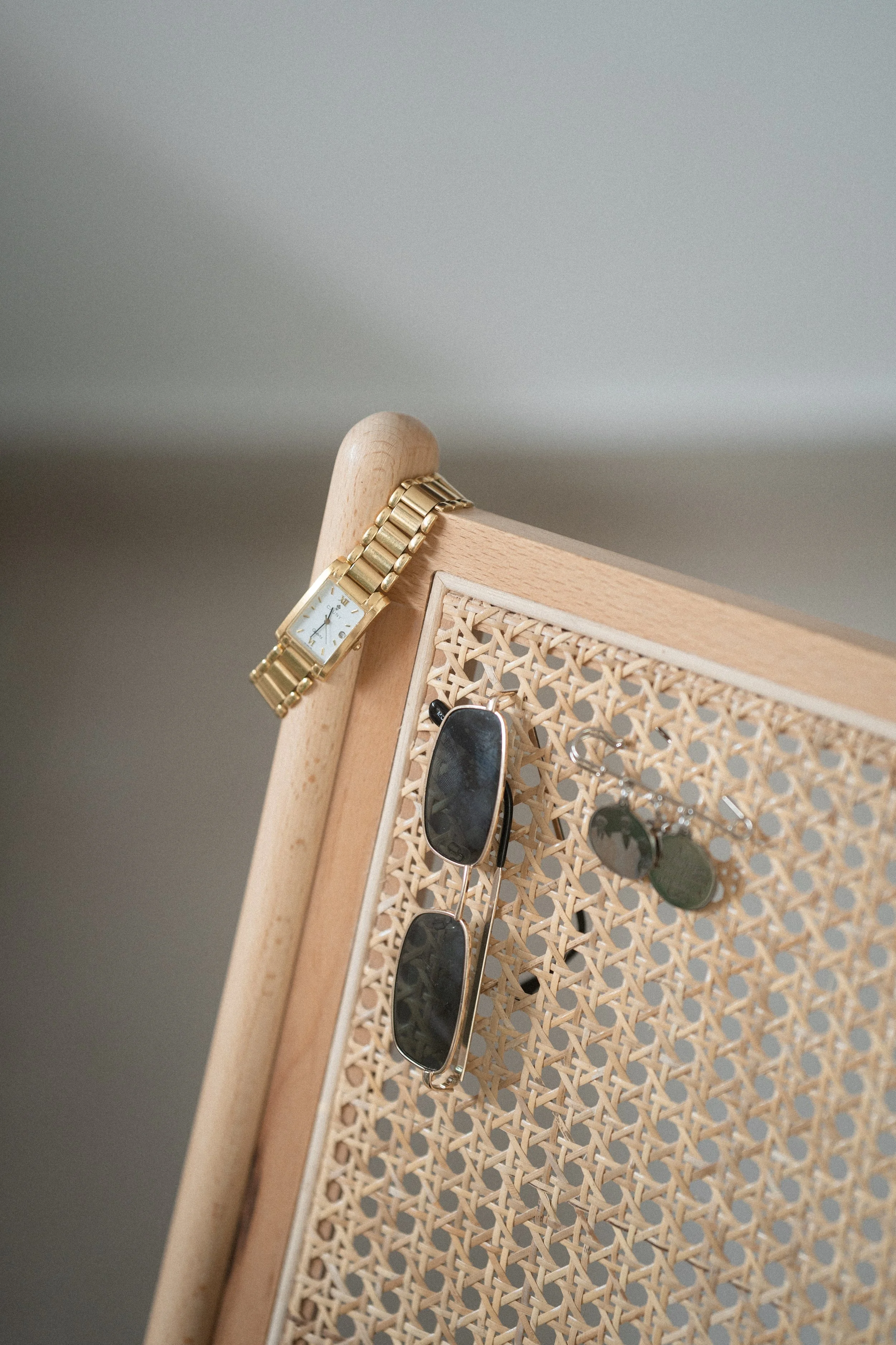 Reloj dorado, gafas de sol con montura metálica, y algunos aretes colgados en una base de caña de madera con un marco de madera clara.