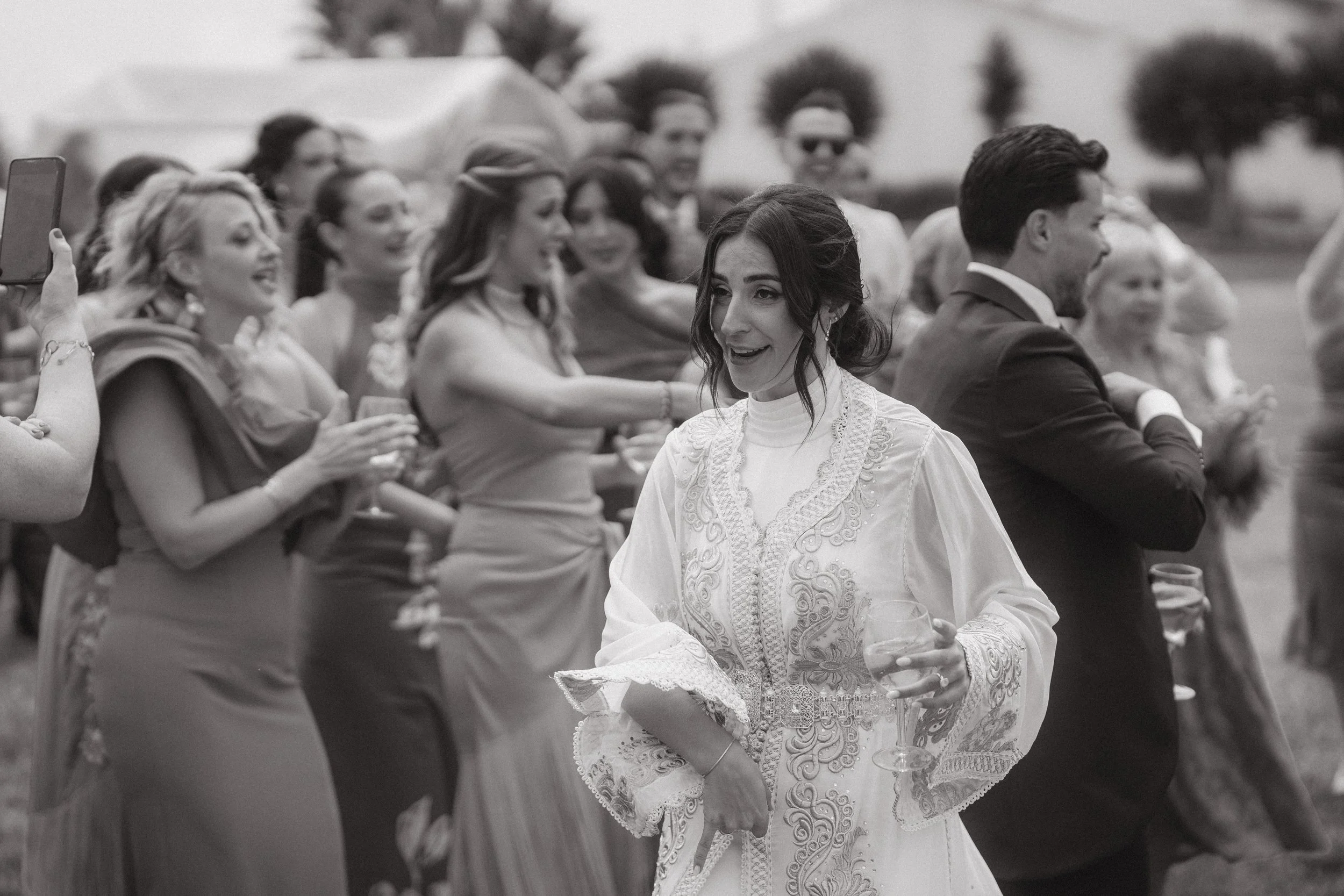 Grupo de personas en una celebración al aire libre, algunas sirven bebidas y disfrutan del evento, con árboles y cielo de fondo.