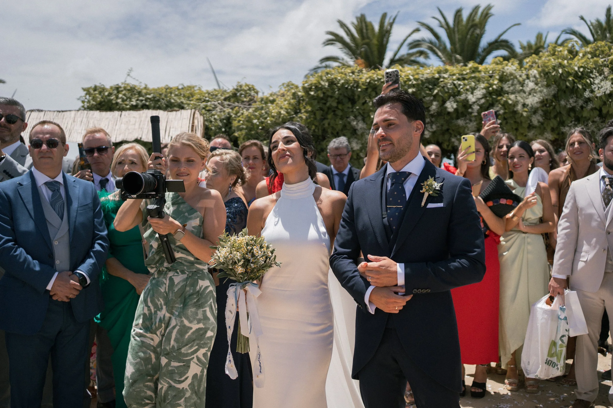 Pareja de novios en una boda, rodeados de invitados, con fondo de palmeras y cielo despejado.