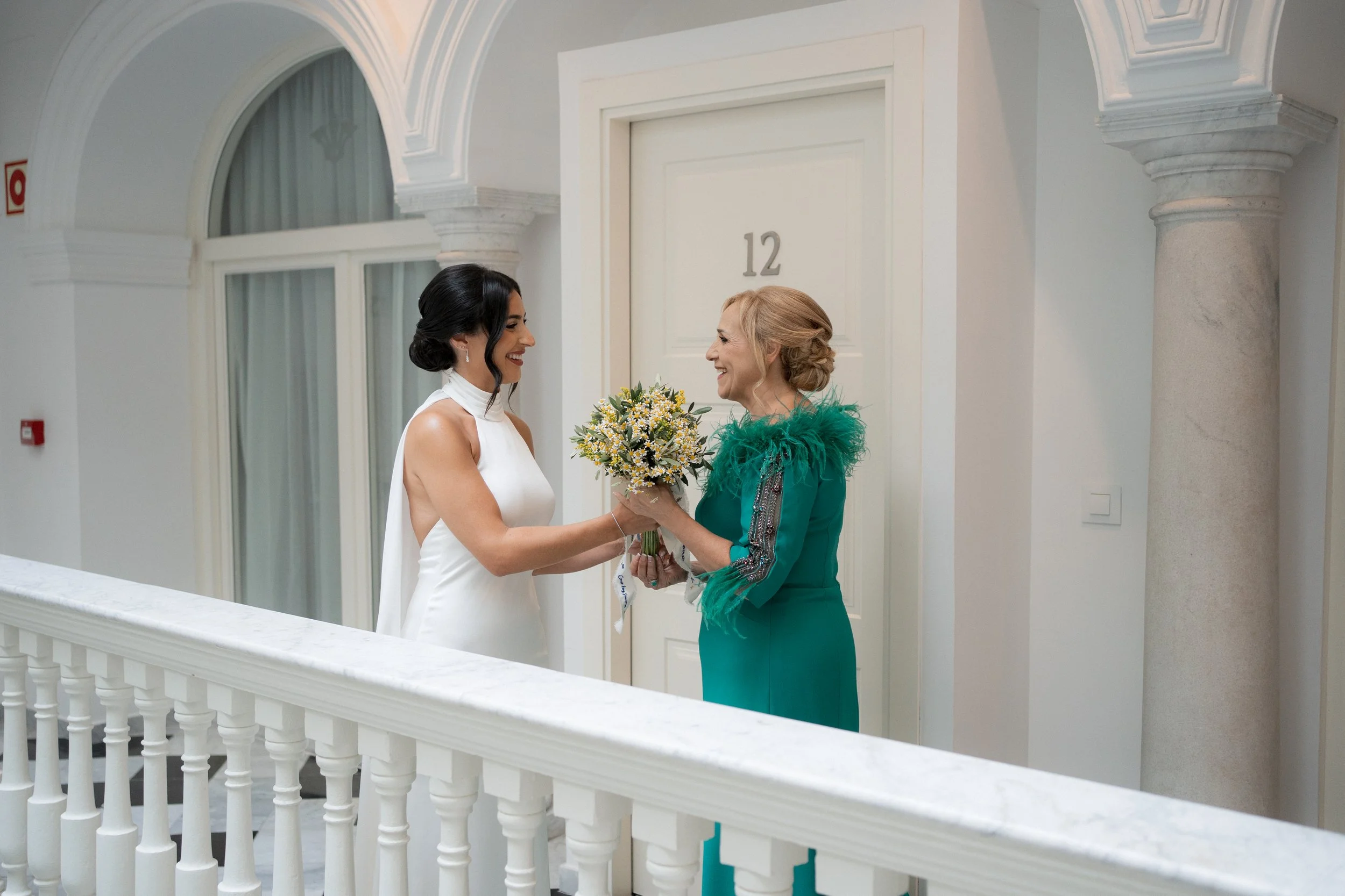 Dos mujeres en un edificio elegante, una con vestido blanco y otra con vestido verde, intercambiando un ramo de flores, ambas sonriendo.