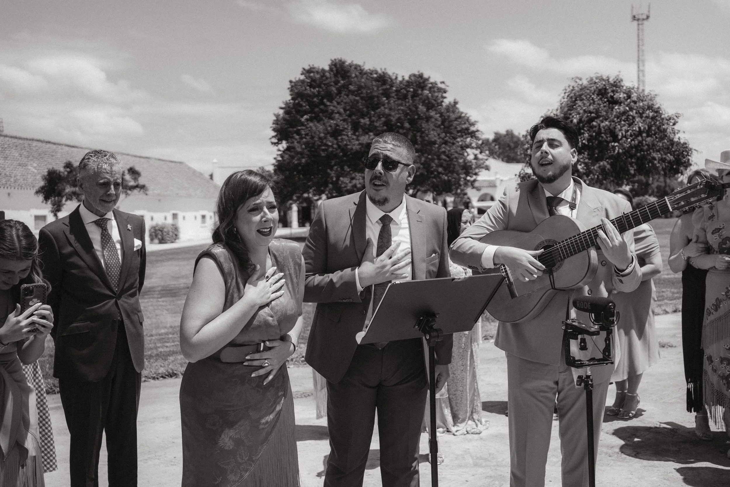 Grupo de personas en una ceremonia al aire libre, algunos cantando y uno tocando la guitarra, con árboles y un cielo con nubes de fondo.