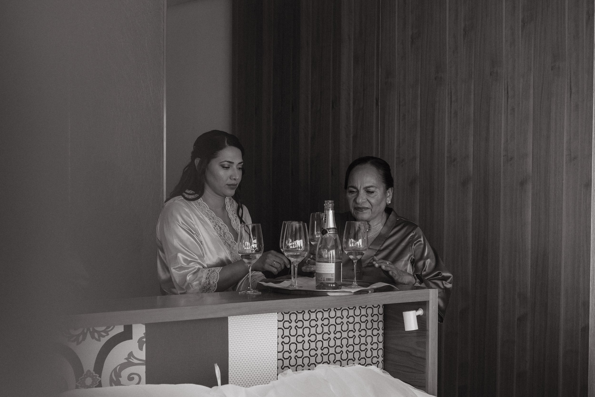 Dos mujeres en batas de satén sentadas en una mesa con copas de vino y una botella, en una habitación con paredes de madera.