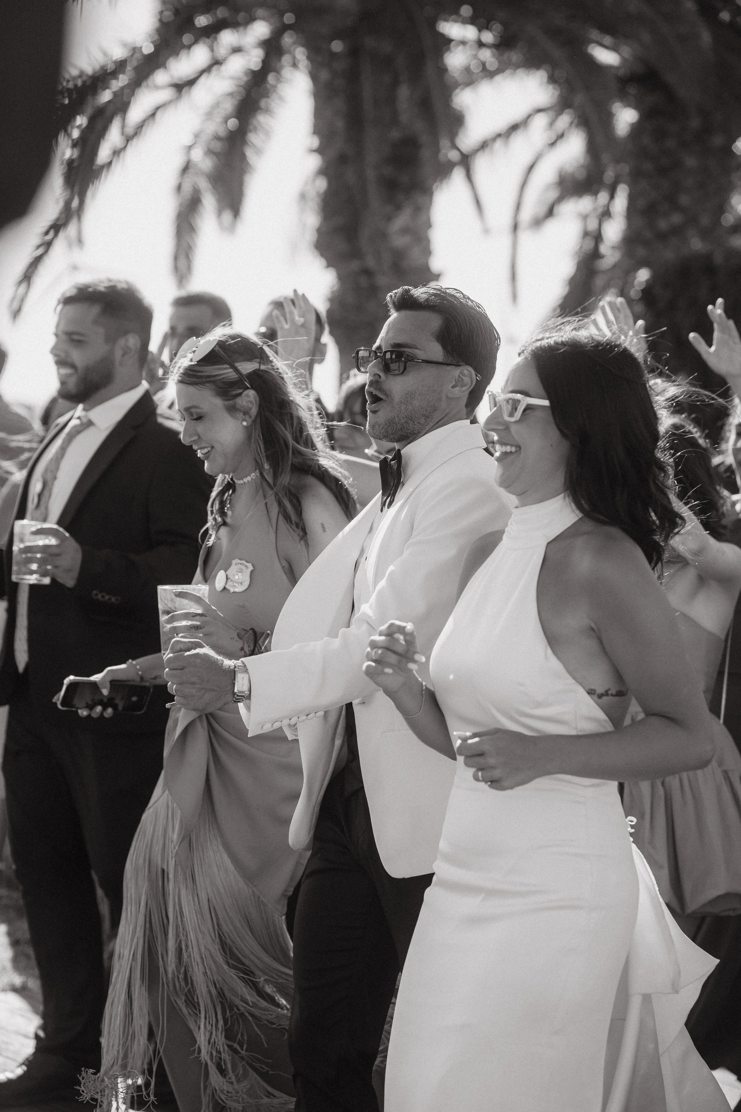Personas en una celebración al aire libre, algunas con gafas, vestido formal y sonriendo.