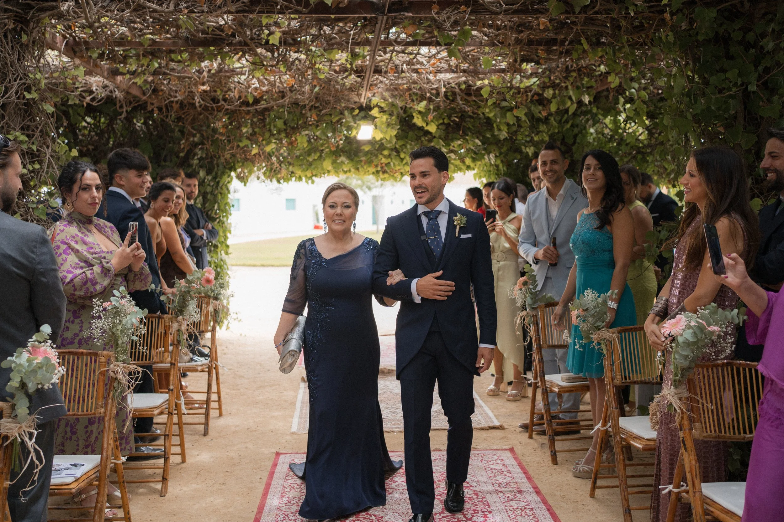 El novio y la madrina entran caminando por pasillo decorado con flores y hojas, rodeados de invitados que toman fotos y observan en un entorno al aire libre con estructura de enredaderas.