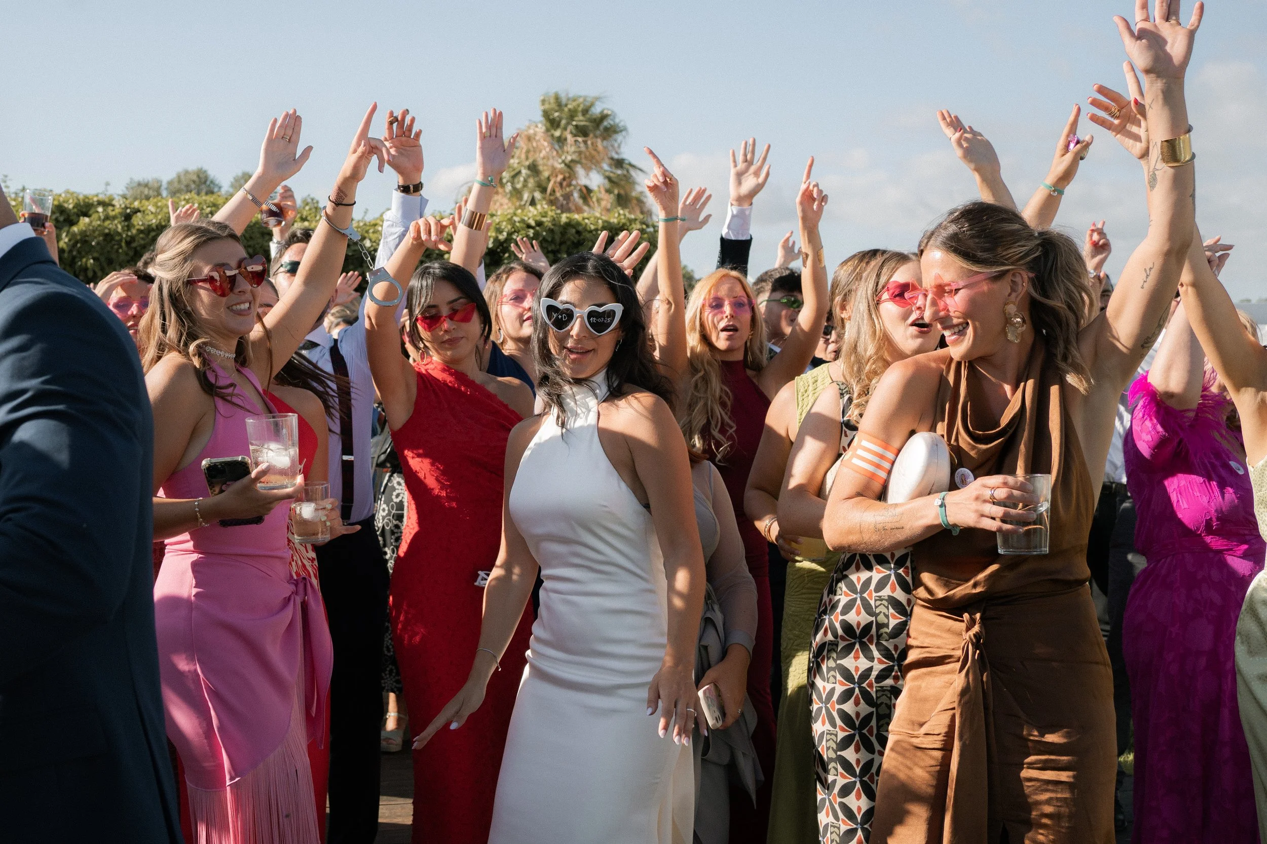 Un grupo de personas en una fiesta al aire libre, algunas con vestidos coloridos y gafas de sol, celebrando con las manos levantadas y sonriendo.