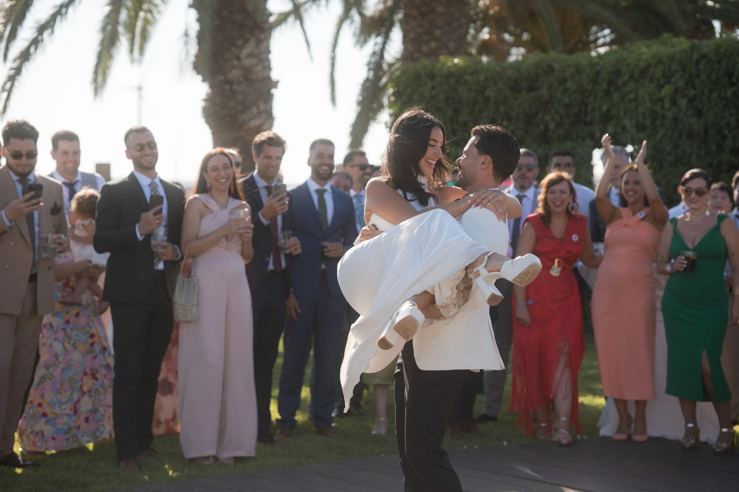 Pareja bailando en una boda al aire libre, rodeados de invitados sonrientes y tomando fotos.
