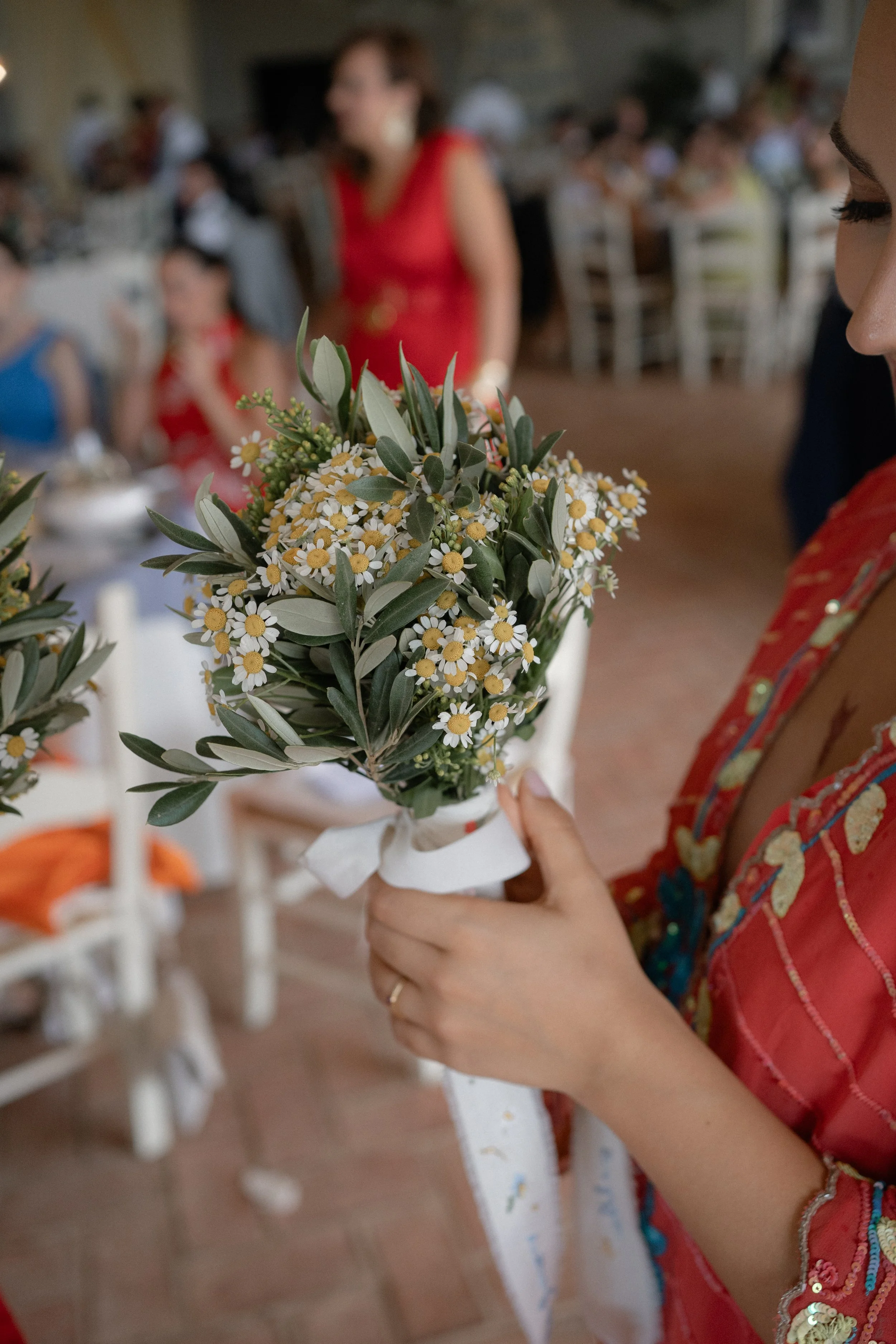 Una mujer sostiene un ramillete de flores de margarita y hojas verdes en una celebración, con personas sentadas y decoraciones en el fondo.