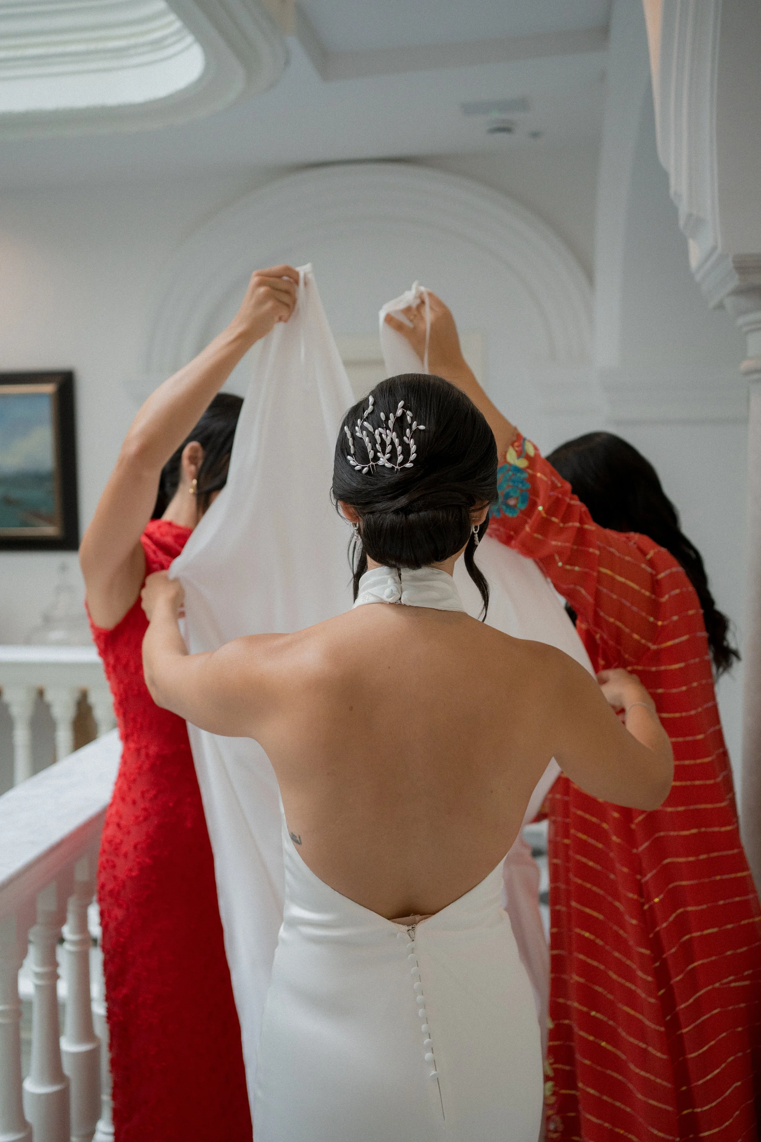 Mujer con vestido de novia ajustando su vestido mientras otras dos mujeres la ayudan en un espacio interior decorado, probablemente antes de una boda.