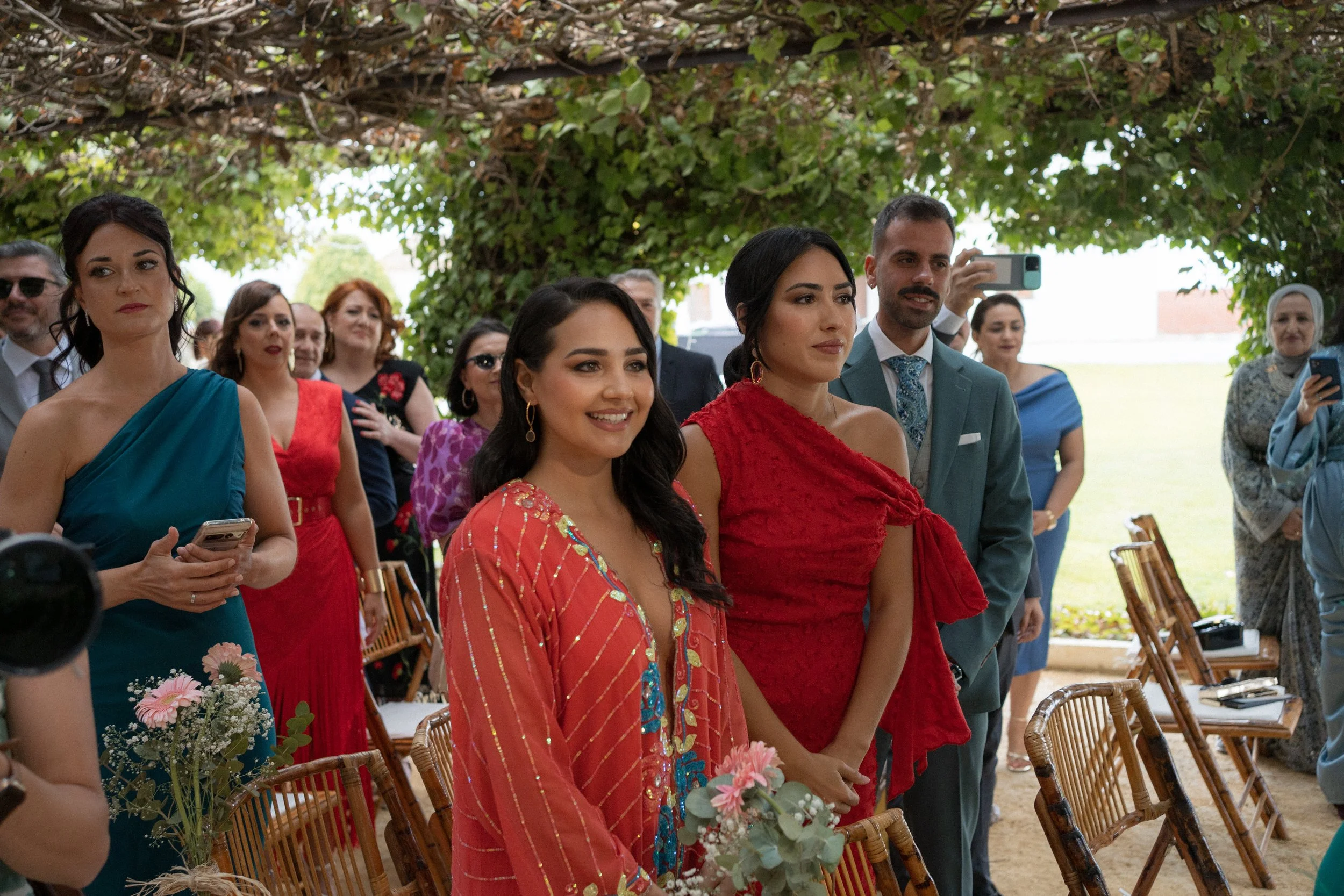 Grupo de personas en una ceremonia al aire libre, incluyendo mujeres con vestidos coloridos y un hombre con traje, bajo un árbol.