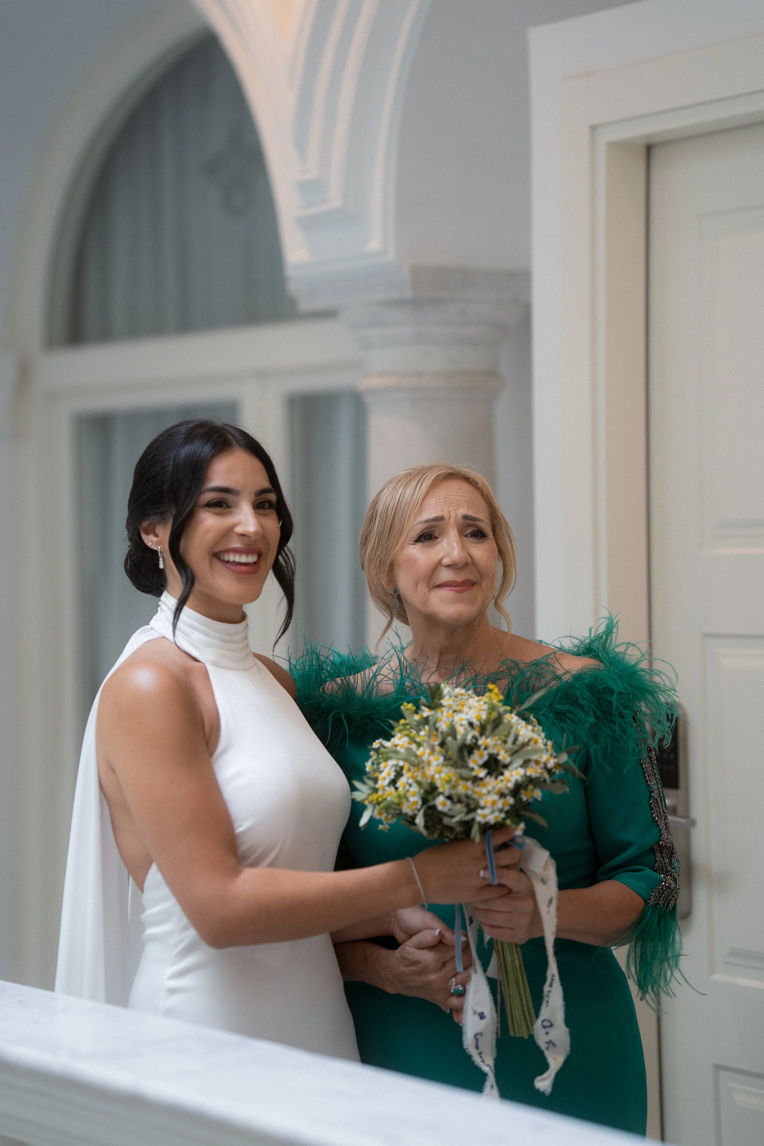 Una mujer joven de peinado recogido y vestido blanco sonriente, junto a una mujer mayor con cabello rubio y vestido azul con detalles de plumas verdes, que sostiene un ramo de flores. Ambas parecen estar en una ceremonia o evento dentro de un lugar e
