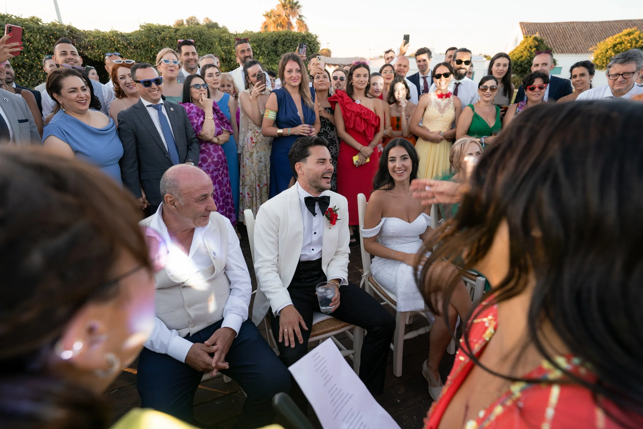 Pareja de novios en matrimonio rodeados de amigos y familiares en una celebración al aire libre, con muchos asistentes sonriendo y tomando fotos.