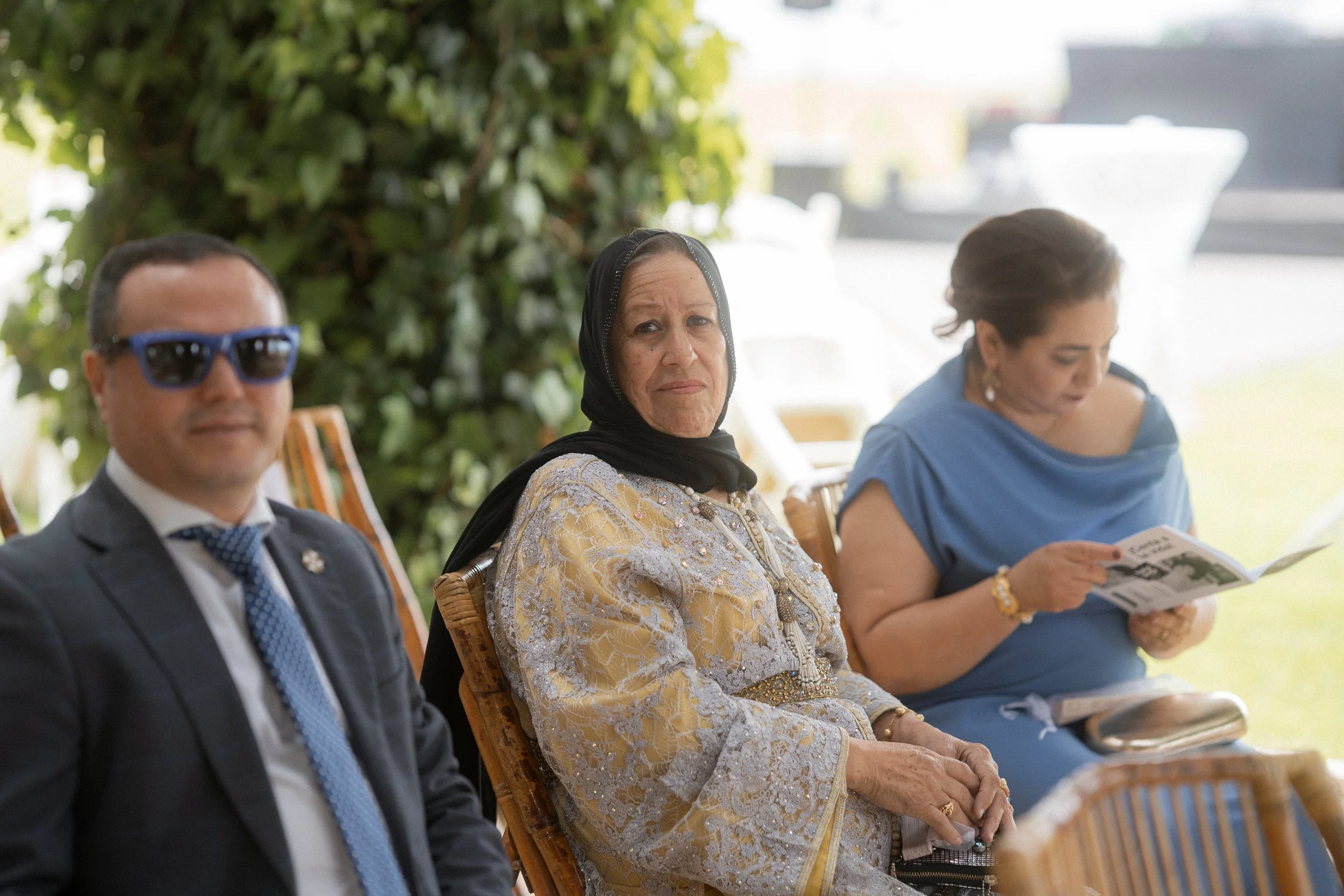 Tres personas sentadas en una reunión, con plantas verdes en el fondo. La mujer del centro lleva un pañuelo negro en la cabeza y un vestido bordado. La mujer a la derecha lee un libro y el hombre a la izquierda lleva gafas y un traje con corbata.
