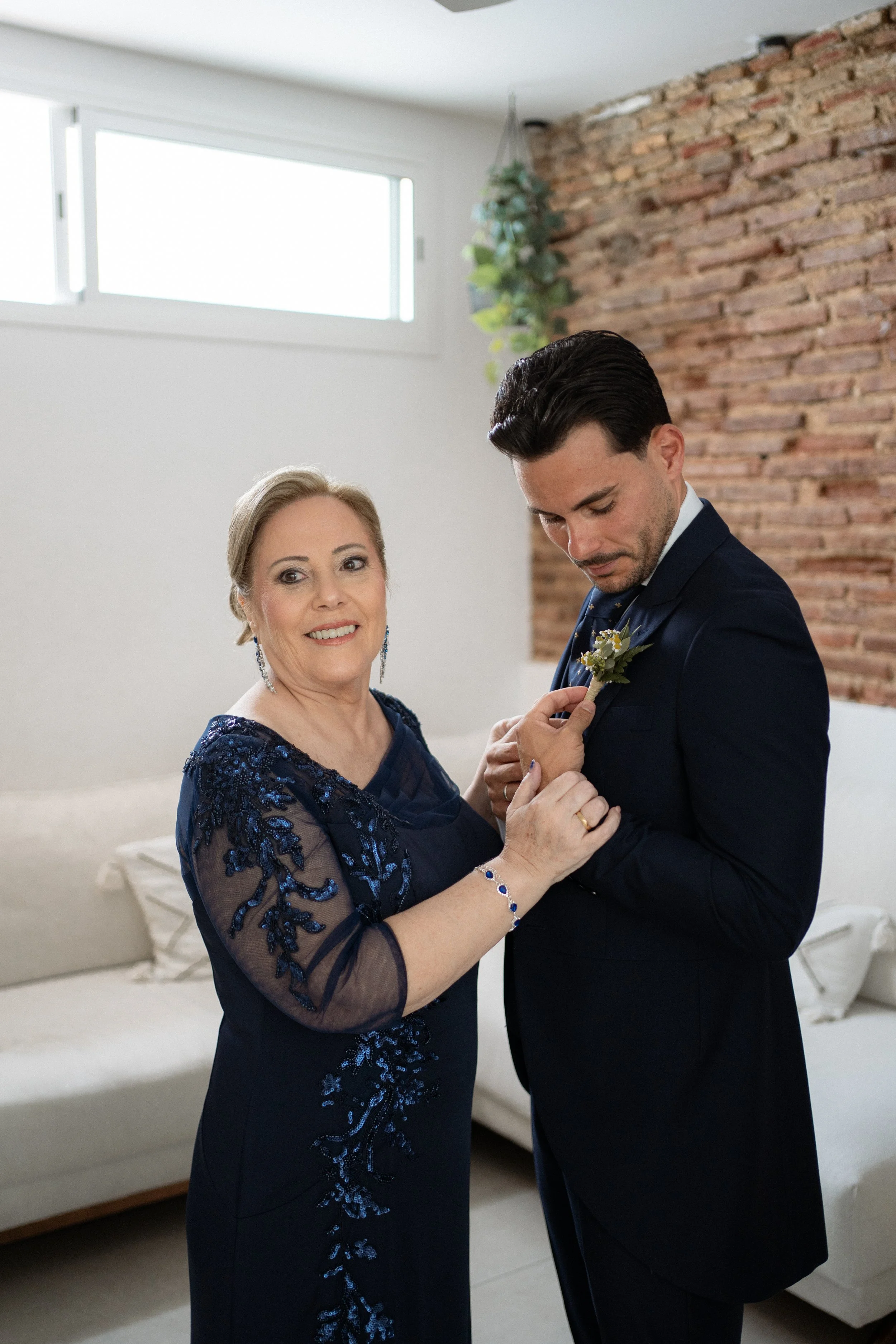 Una mujer mayor en un vestido azul con detalles de encaje ayuda a un hombre joven a ponerse una flor en su chaqueta, en un ambiente interior con pared de ladrillos y sofá beige.