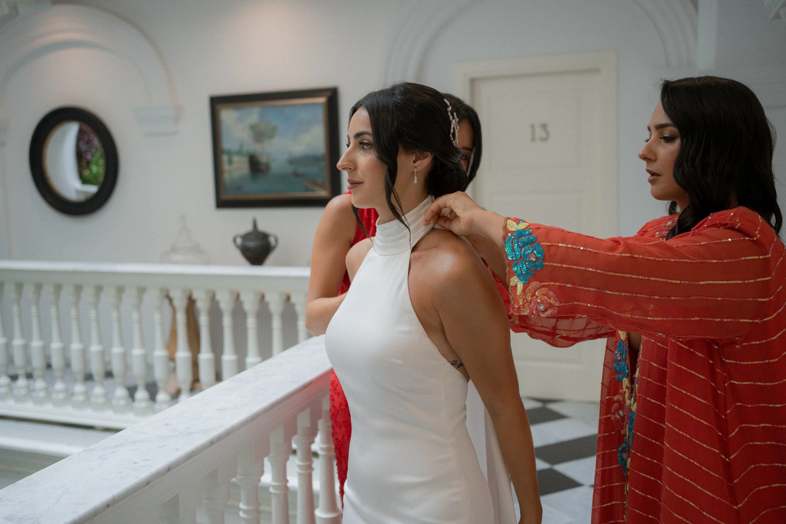 Novia en vestido blanco siendo ajustada para la ceremonia por otra mujer con vestido rojo con bordados azules y dorados.