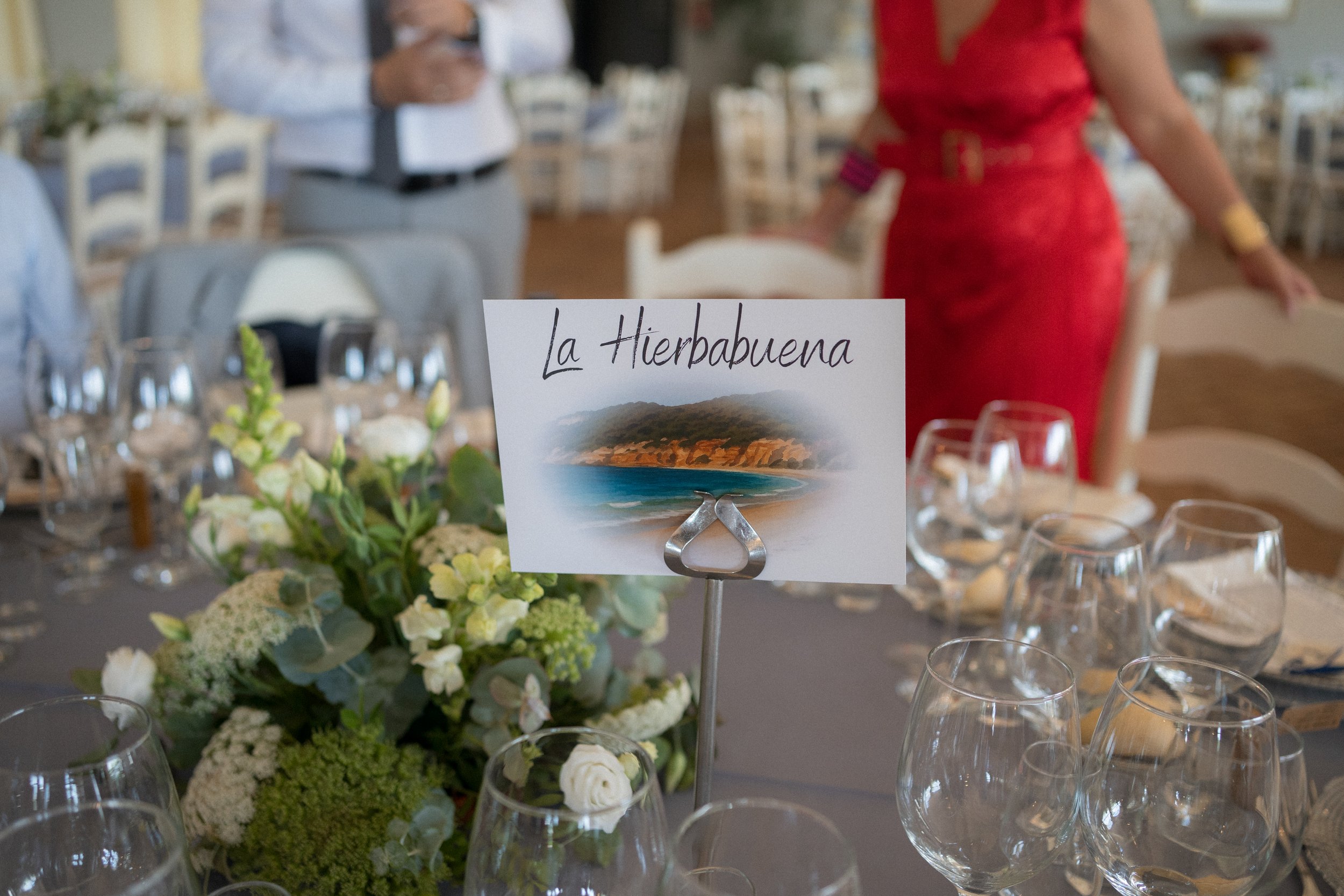 Centro de mesa con flores y cartel que dice "La Hierbabuena" en una mesa de boda, con varias copas y platos.