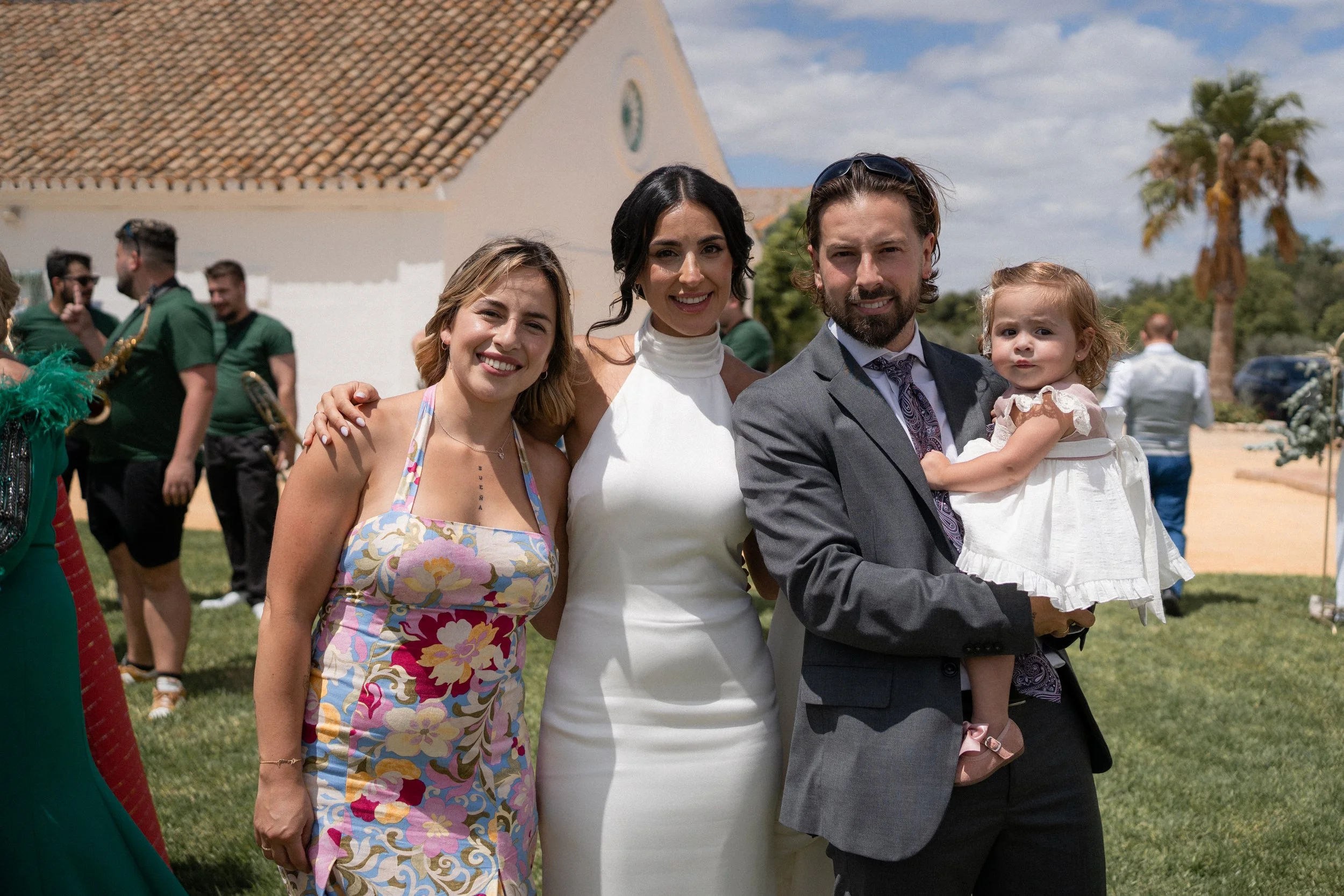 Grupo de personas en una celebración al aire libre, incluyendo un hombre con traje y una niña en brazos, una mujer con vestido blanco y dos mujeres con ropa colorida, en un día soleado con árboles y cielo parcialmente nublado en el fondo.