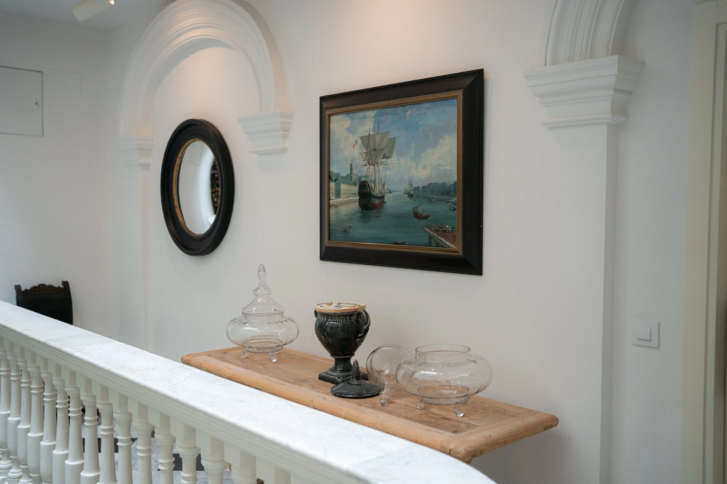 Interior de una estancia con paredes blancas, decorada con un cuadro de un barco y un espejo ovalado, con una repisa de madera que sostiene recipientes de vidrio.