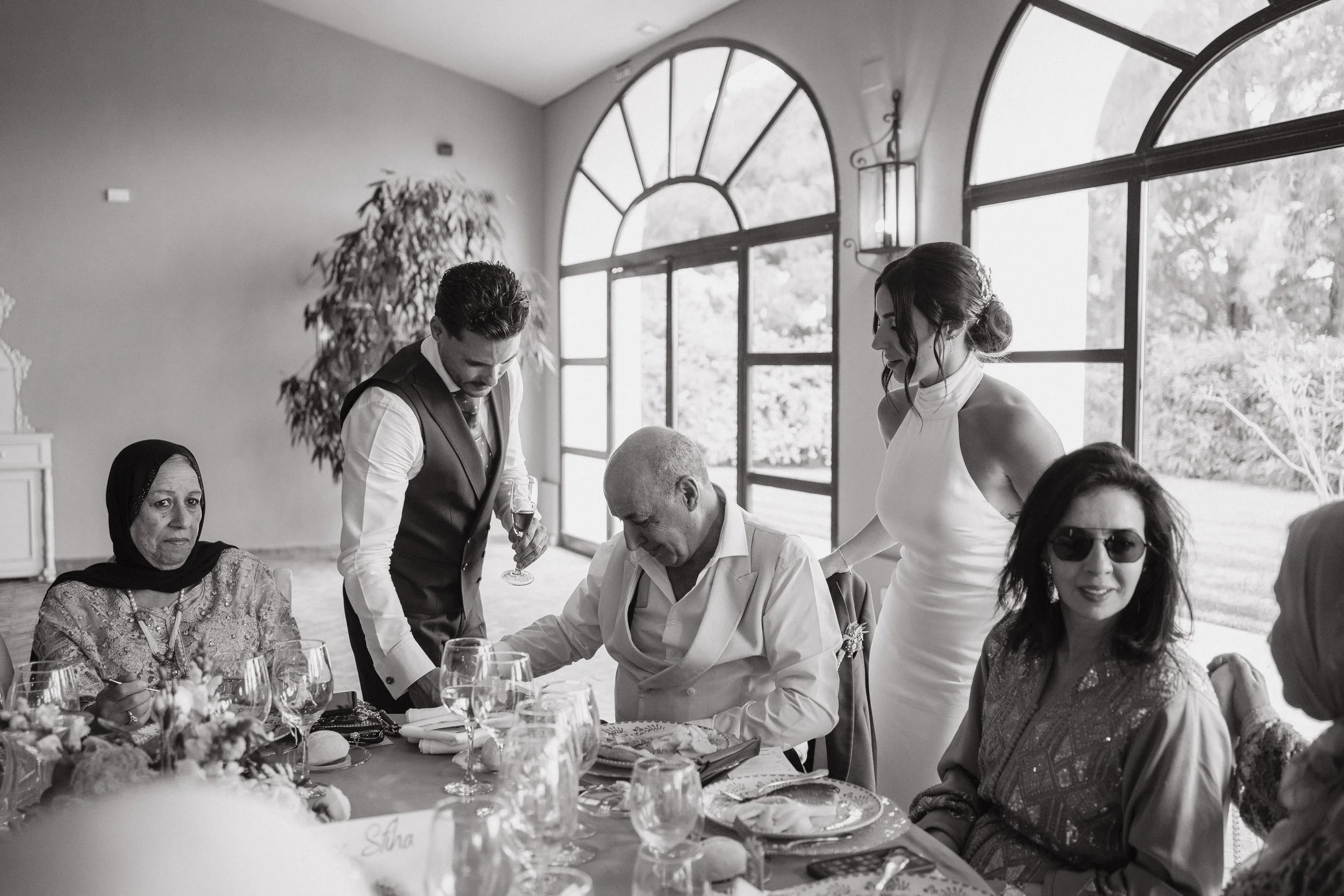 Personas en un banquete de bodas con grandes ventanas, una de ellas con arco, y decoración elegante, en un ambiente luminosa y relajado.