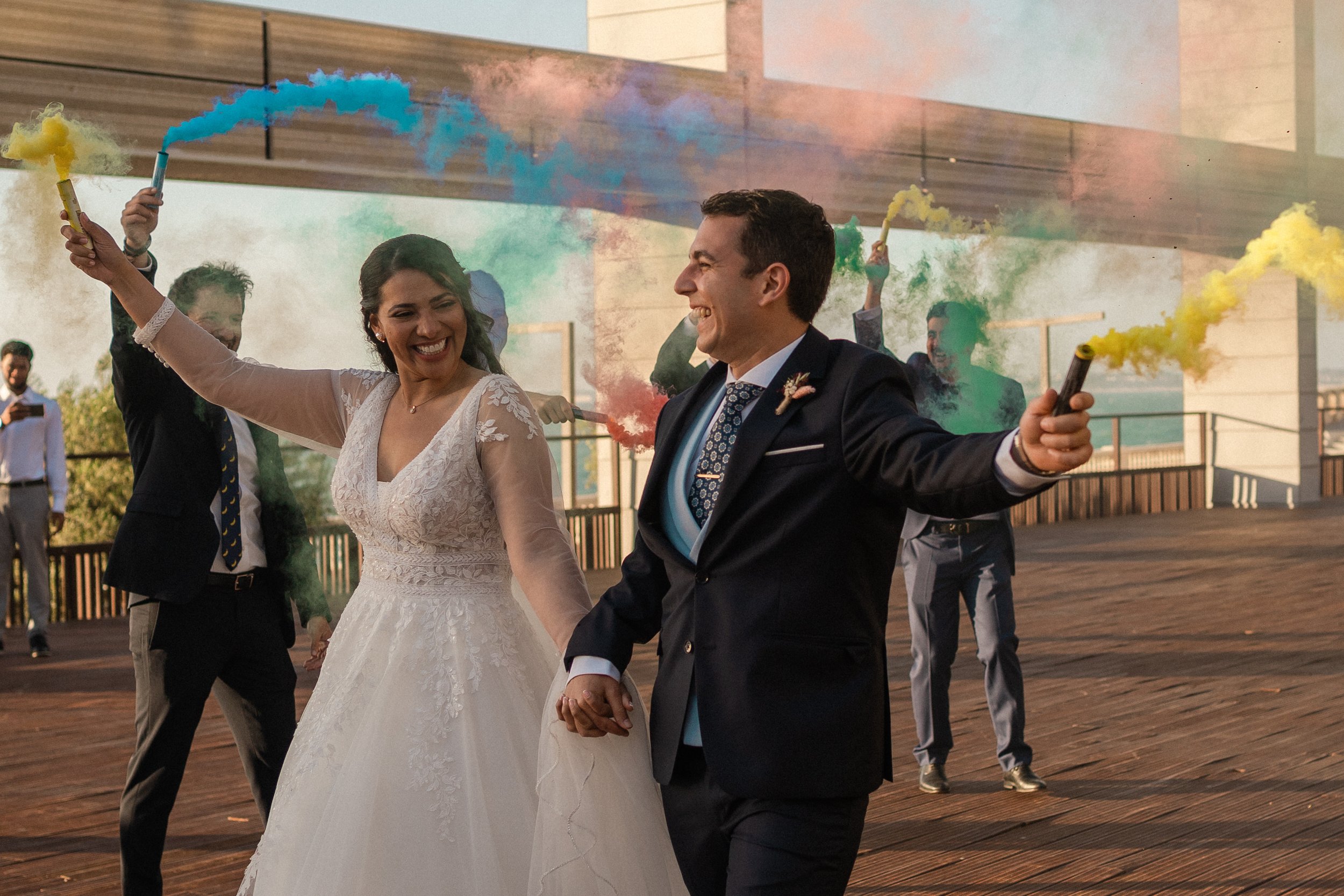 Pareja de novios caminando en una celebración de boda, rodeados de invitados sosteniendo bengalas de humo de colores. El ambiente es festivo y alegre.