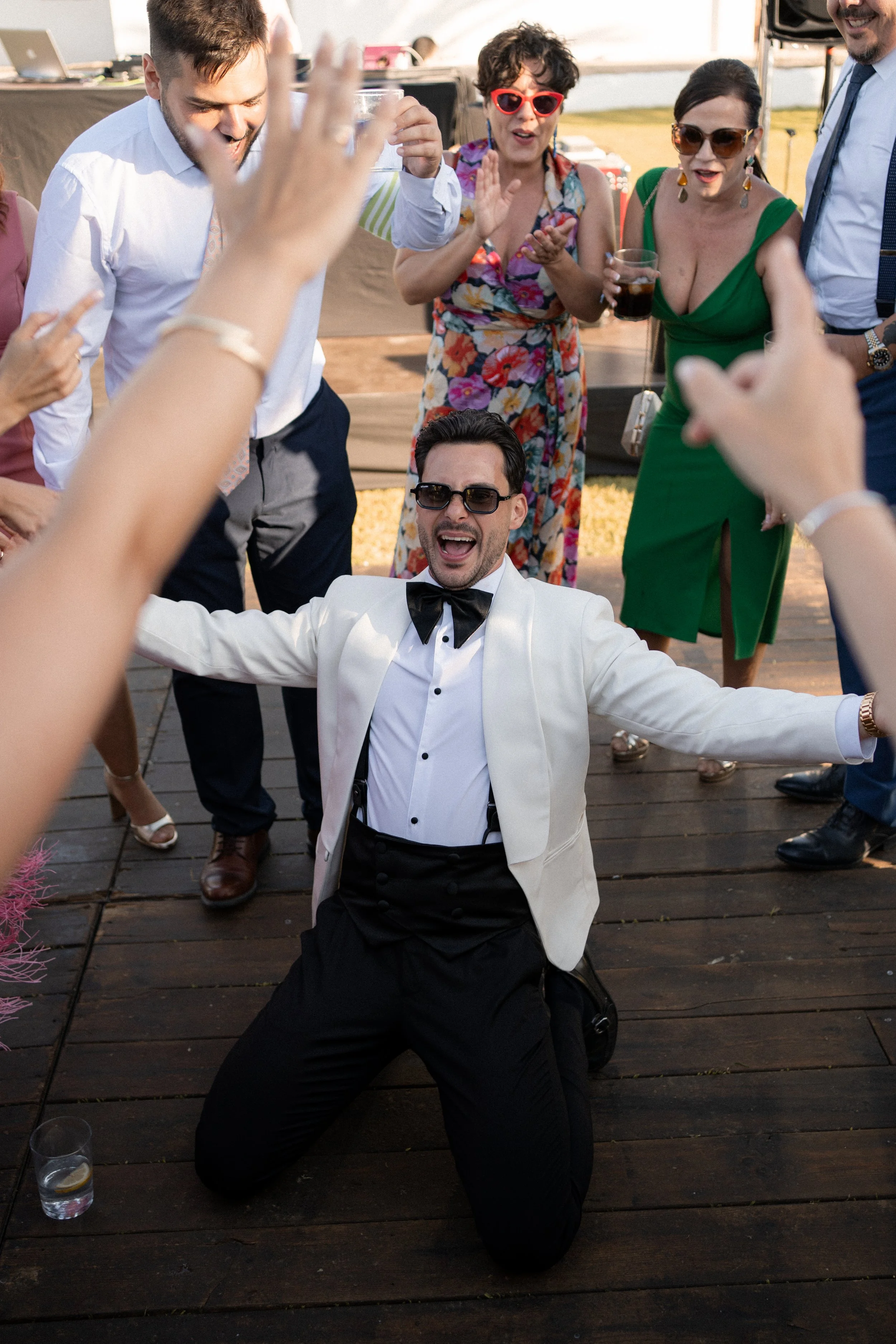 Hombre vestido de esmoquin blanco con pajarita negra, con gafas negras, arrodillado en el suelo con los brazos abiertos, rodeado de personas que bailan y se divierten en una celebración al aire libre.