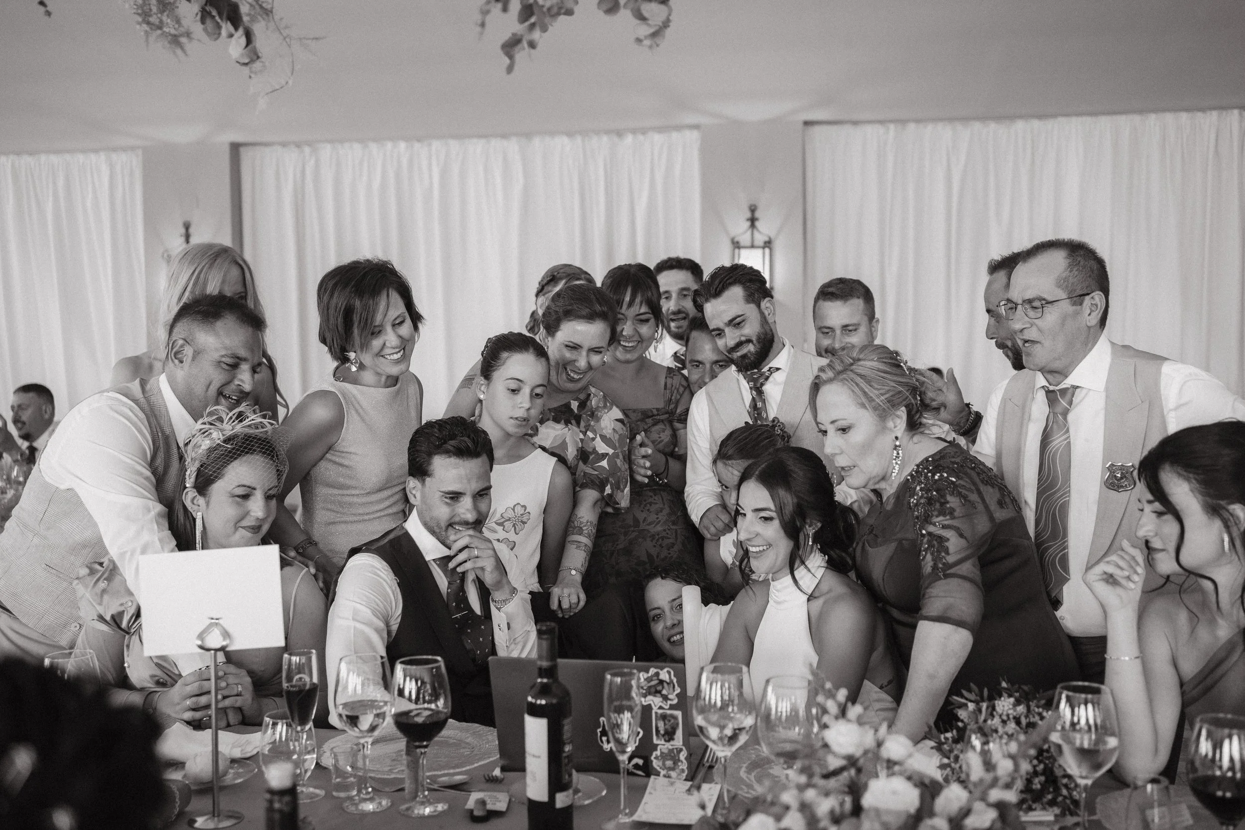 Grupo de personas en celebración, mirando una pantalla en una mesa con comida y bebidas, en un evento social o boda, en blanco y negro.