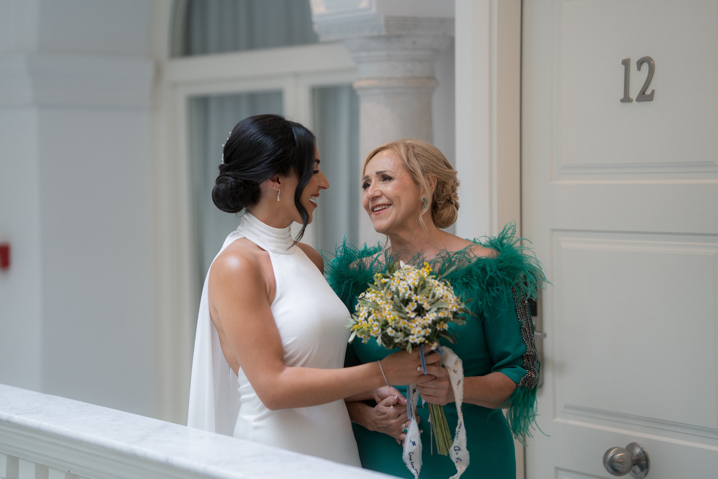 Una joven novia sonriendo y hablando con su madre, que le da un ramo de flores, frente a una puerta del hotel armador en cadiz.