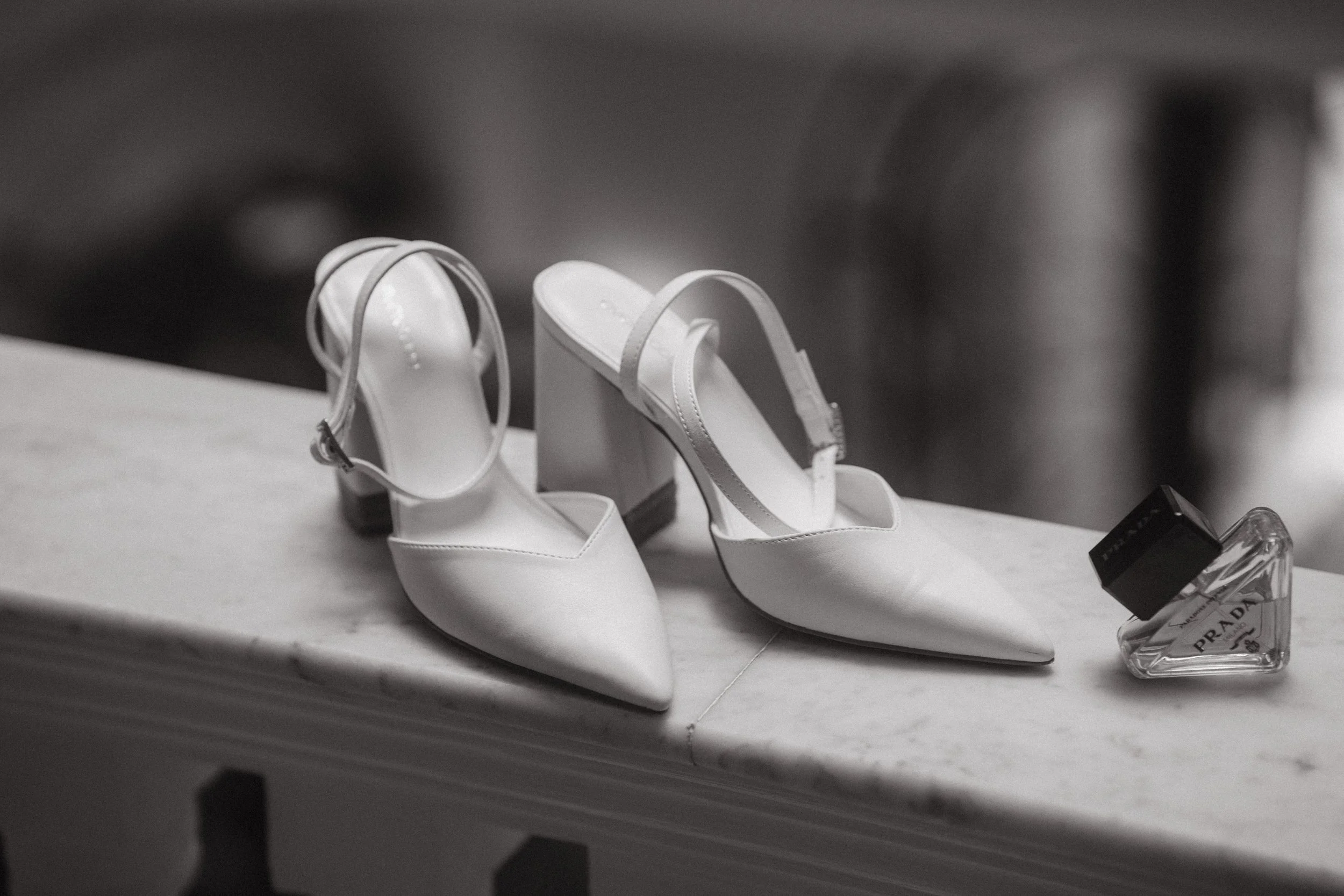 Zapatos de tacón blancos y perfume Prada en una mesa de mármol.